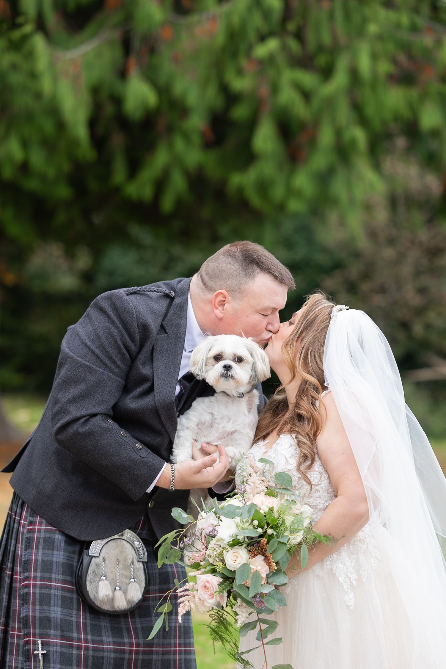 A wedding couple shares a tender moment while holding their small white dog in formal attire outdoors. Huntingtower Hotel