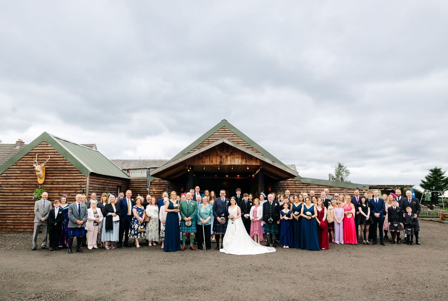 Large wedding group photo of bride, groom, and guests posing outside a rustic barn venue under cloudy skies.
