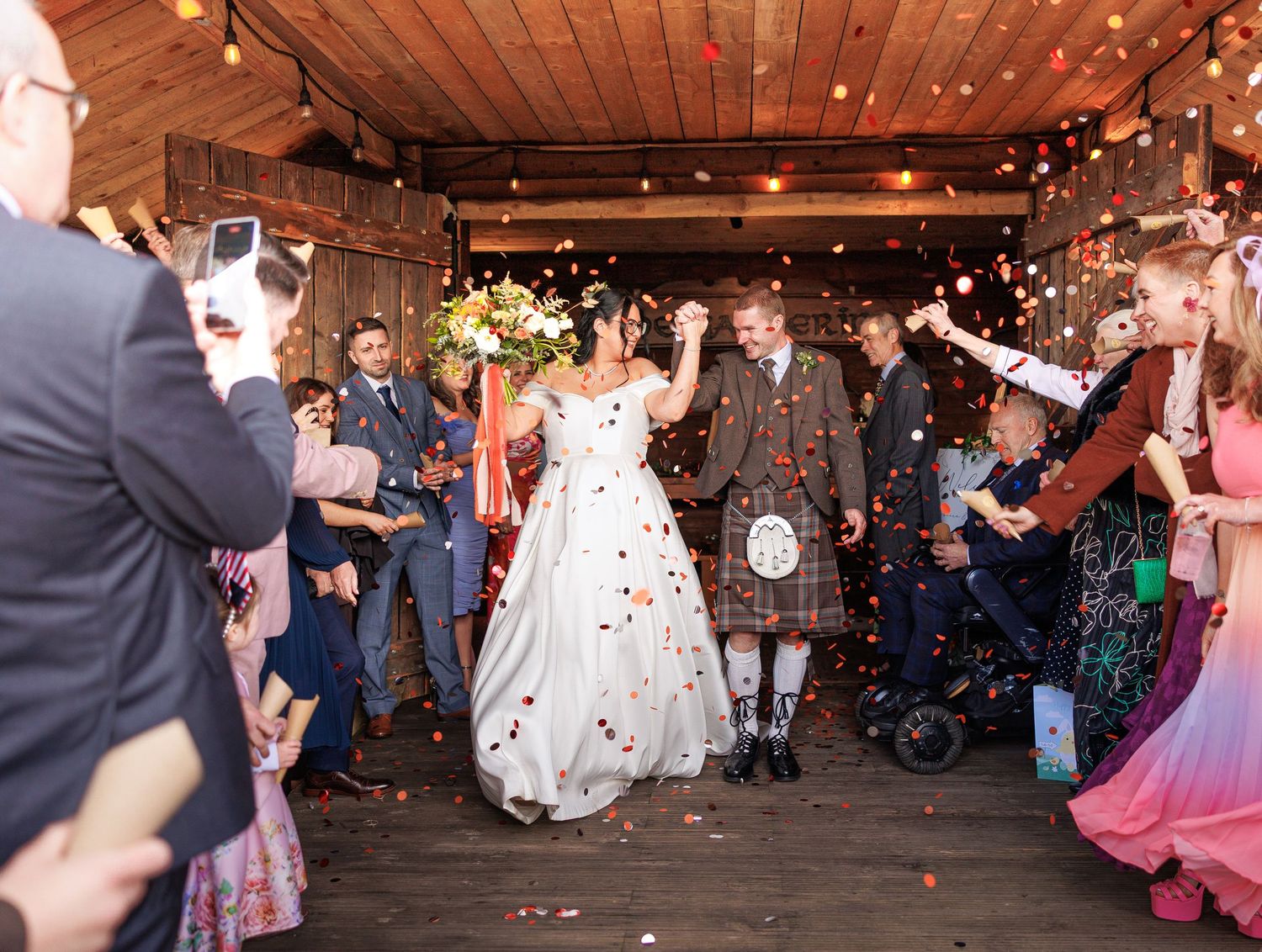 Wedding guests dance and celebrate on a rustic wooden dance floor under twinkling lights at an indoor reception.
