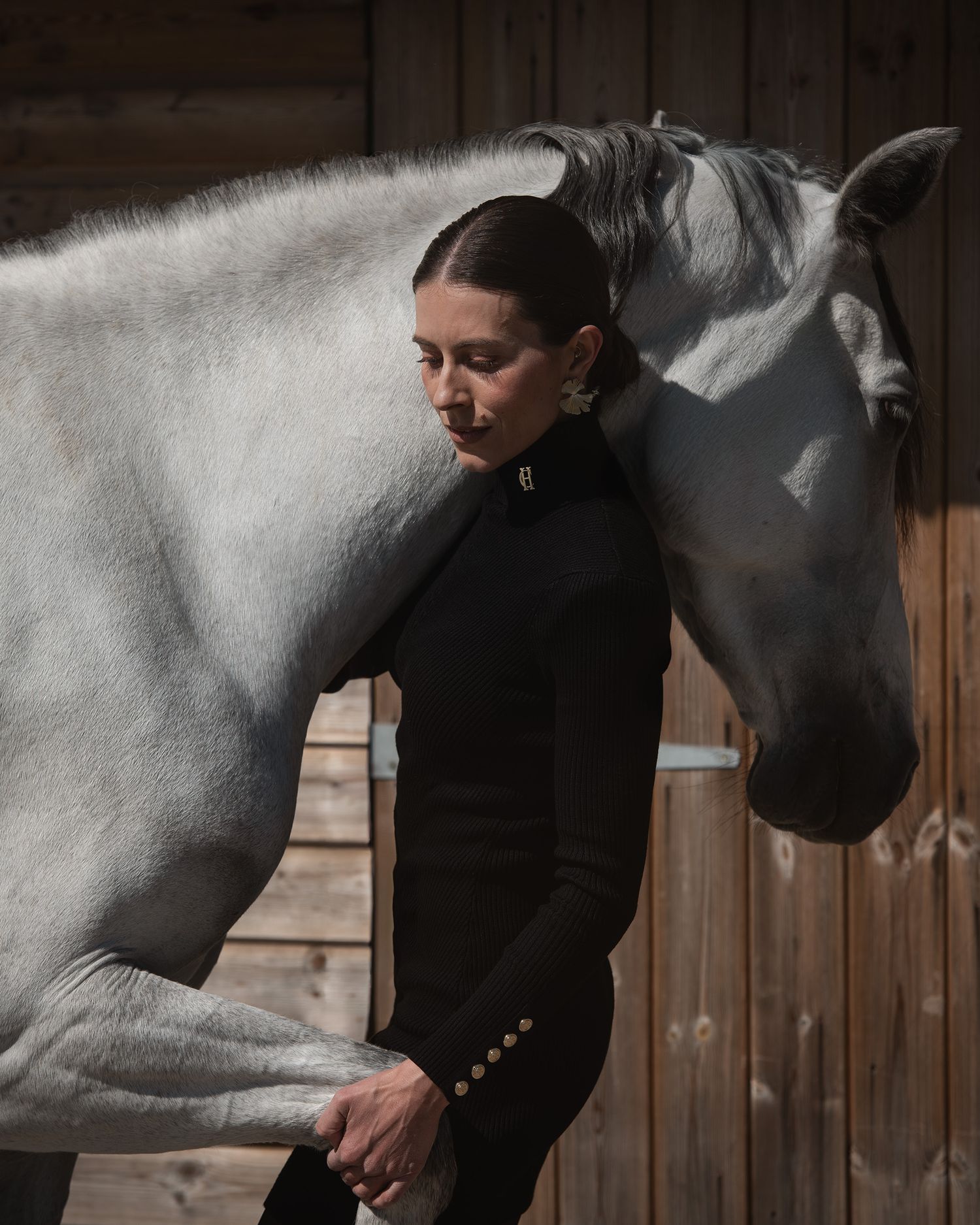 Woman in black equestrian attire stands beside a grey horse in a stable, sharing a serene moment.