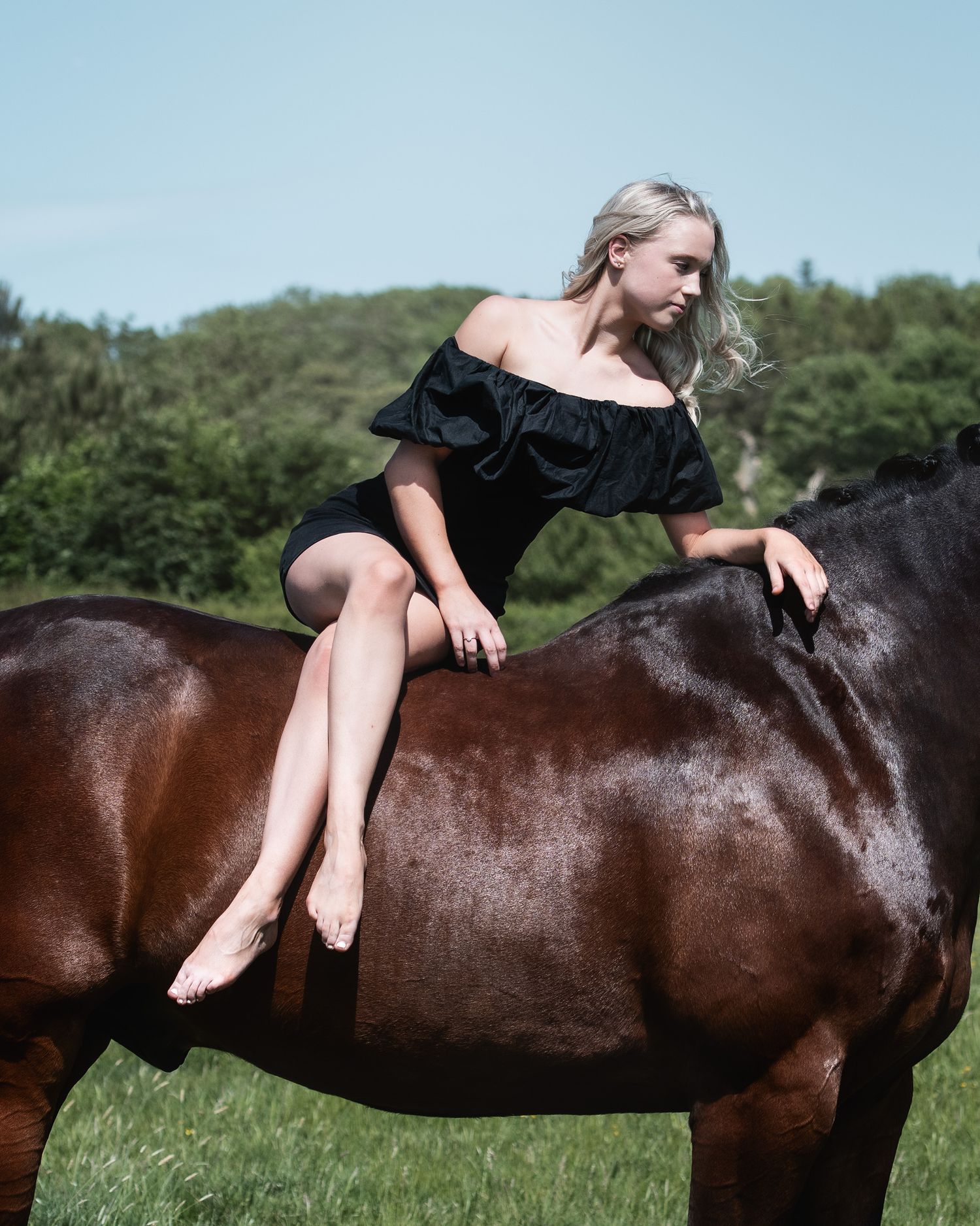 Blonde woman in black off-shoulder dress sitting barefoot on a brown horse in a green field.
