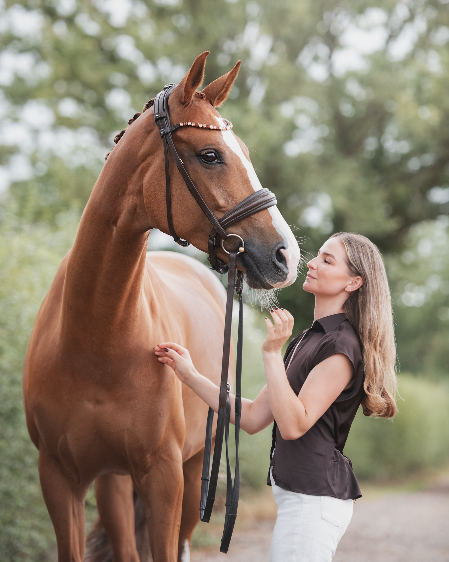 A woman in a dark top and white pants affectionately interacts with a chestnut horse wearing a decorated bridle outdoors.