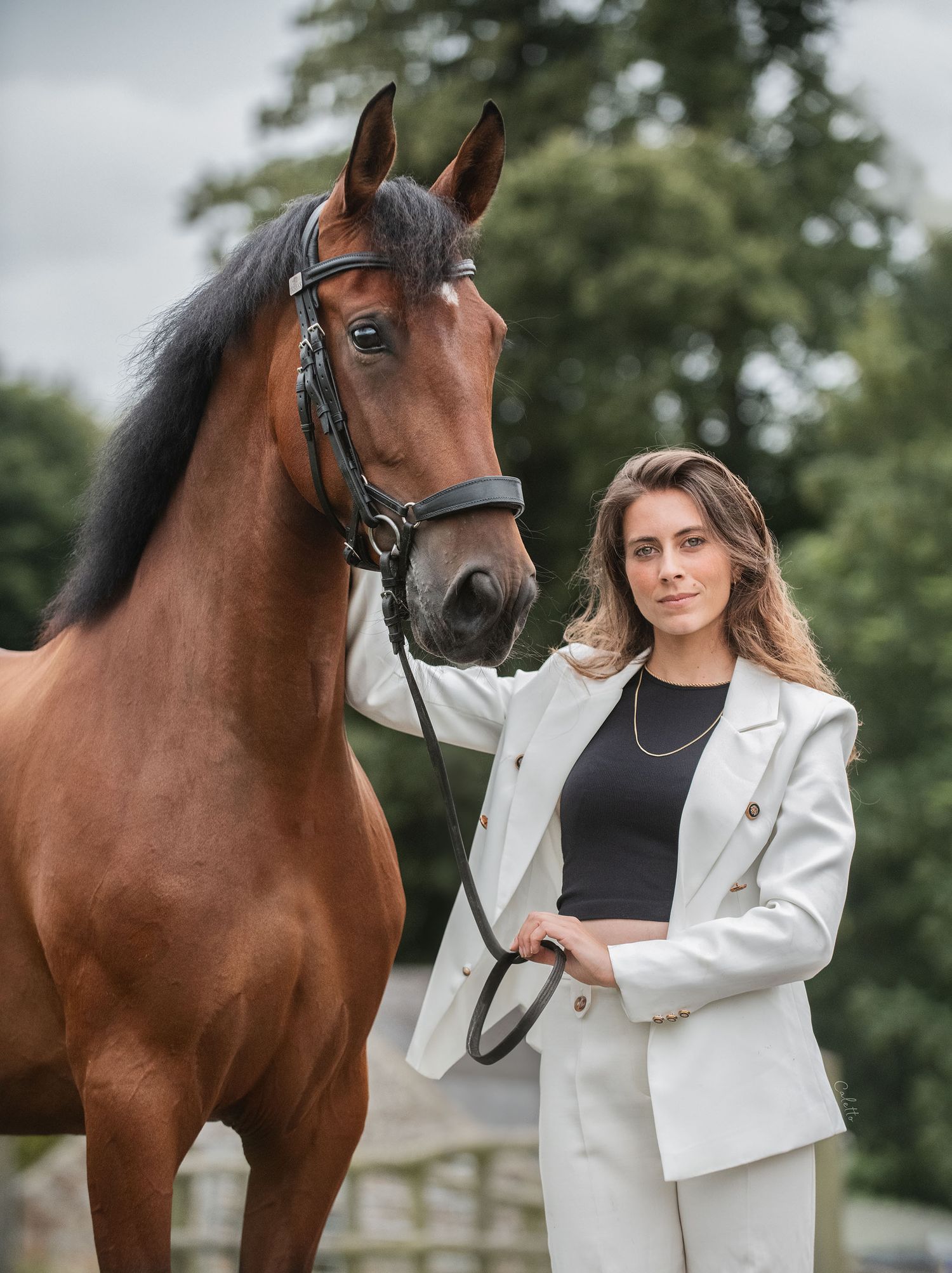 Elegant bay horse wearing a bridle poses outdoors with a white-suited cs equine model & PSG dressage coach Andrea Baliarda against foliage.