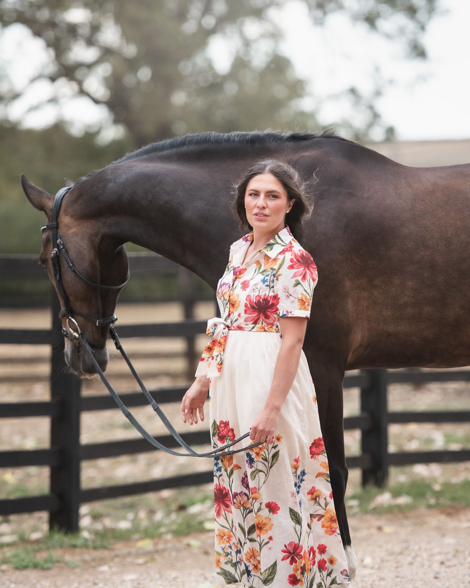 Woman in floral dress standing with dark brown horse in equestrian paddock setting.