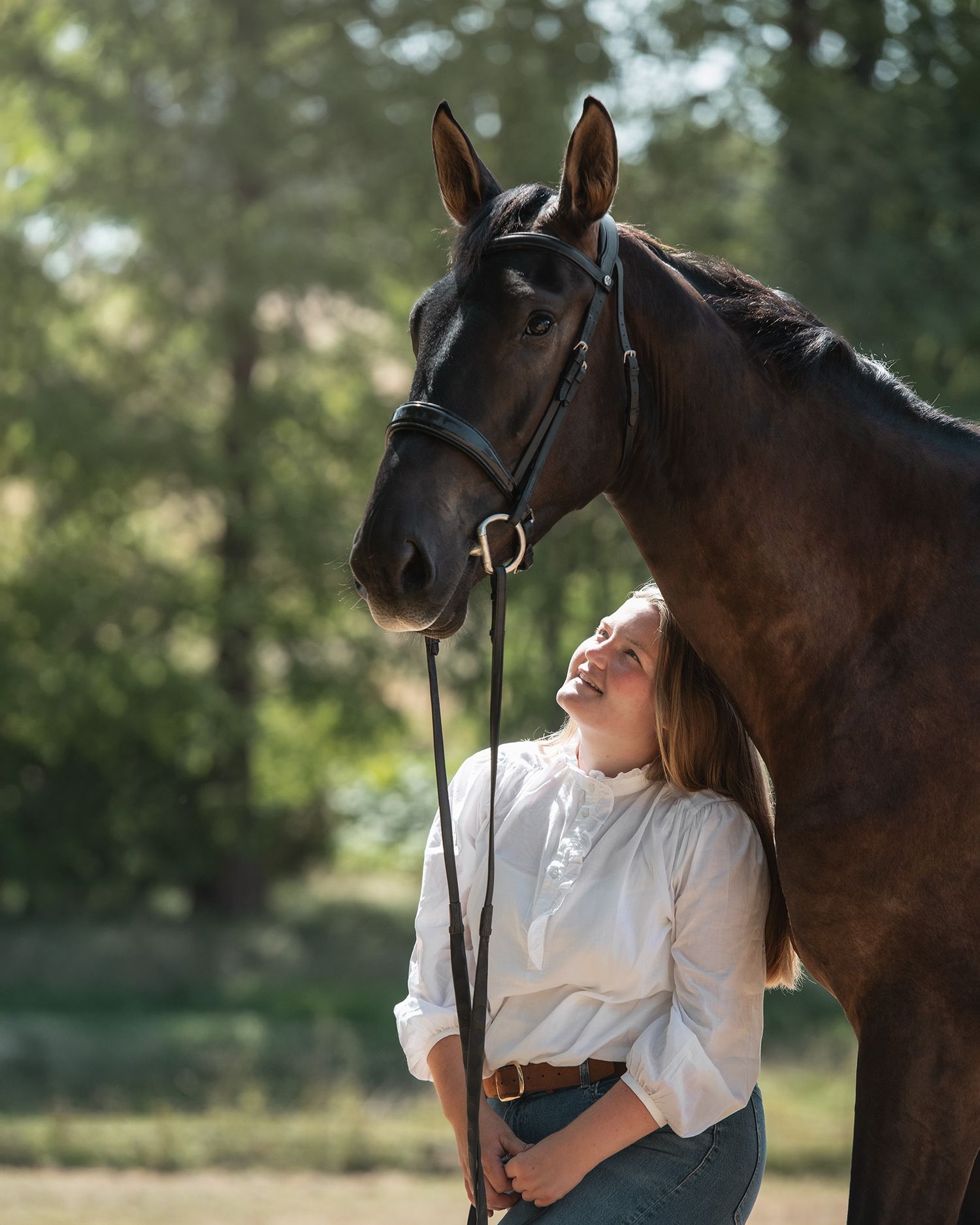 Young woman in white blouse smiling beside a dark brown horse on a lead rope outdoors.