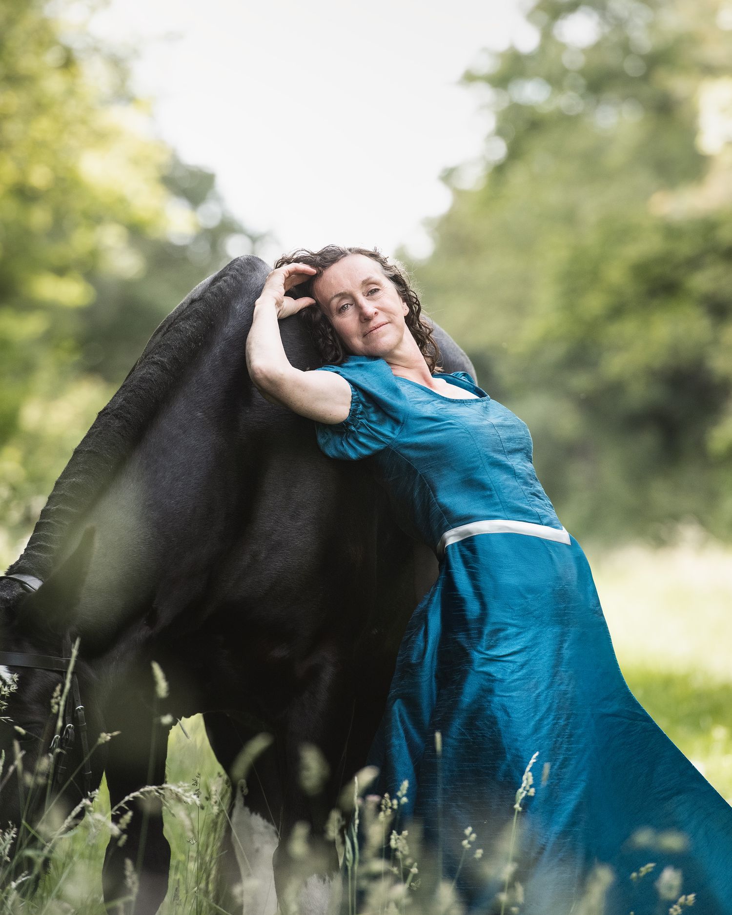 Woman in teal dress embracing a black cow in a lush green meadow, sharing a tender moment with the animal.