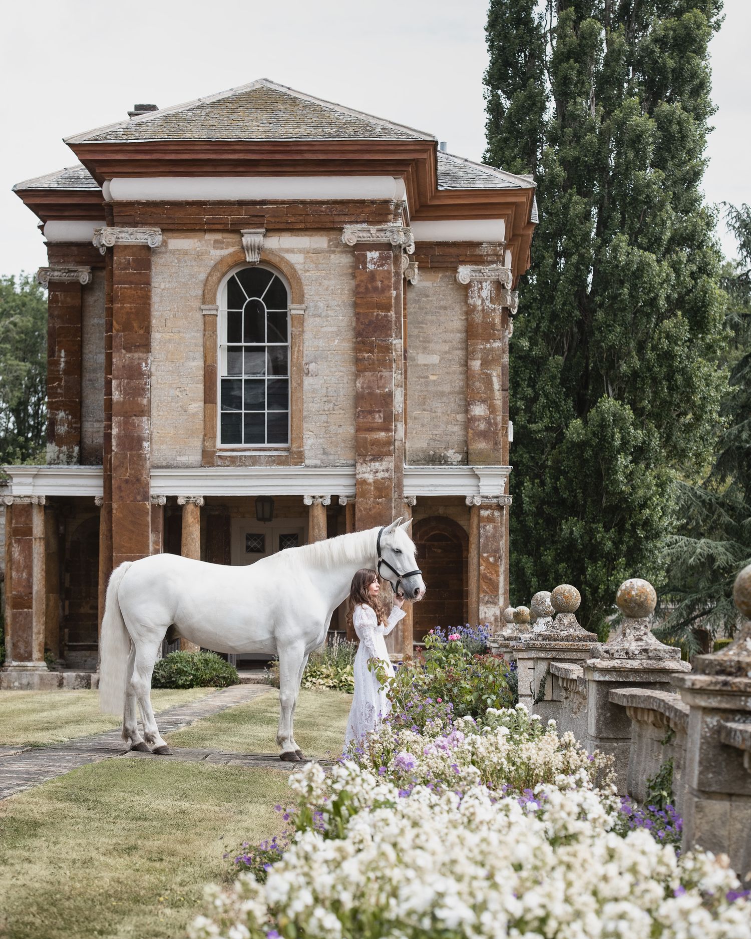 Woman in white dress stands beside a white horse near a grand brick manor house with tall cypress trees.