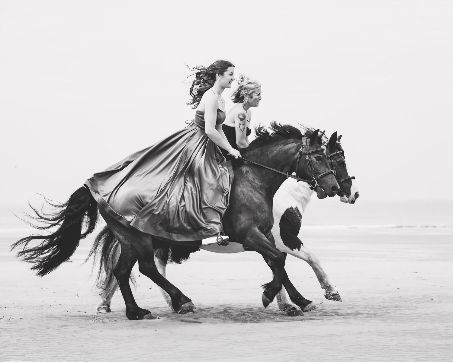 Two women in elegant dresses ride a galloping horse on a beach in a stunning black and white photograph.