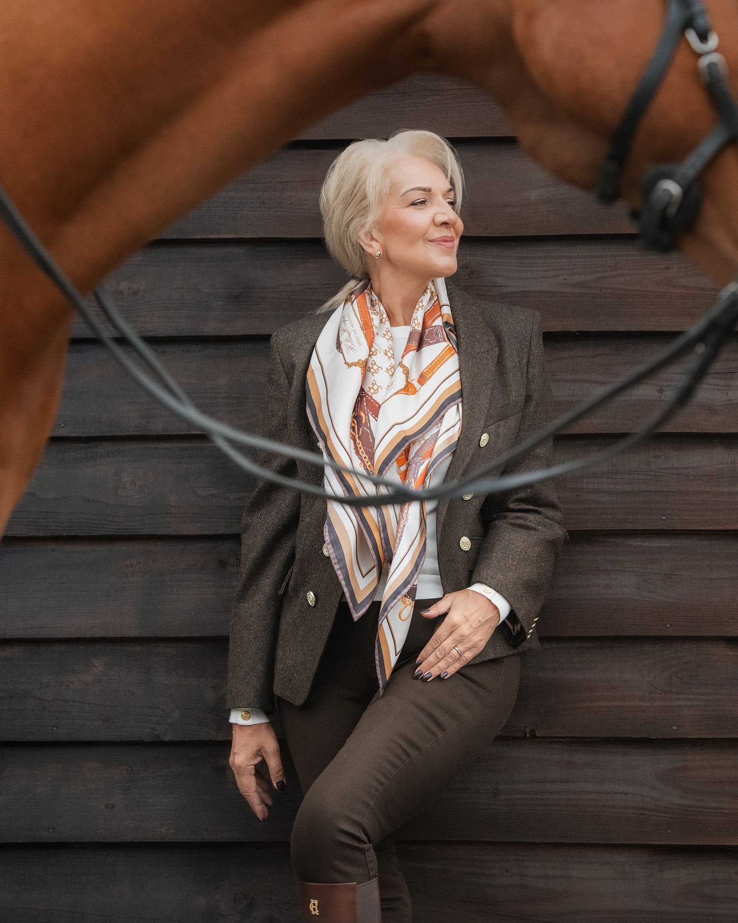 Elegant older woman in equestrian style outfit with silk scarf and blazer posing near a horse stable.