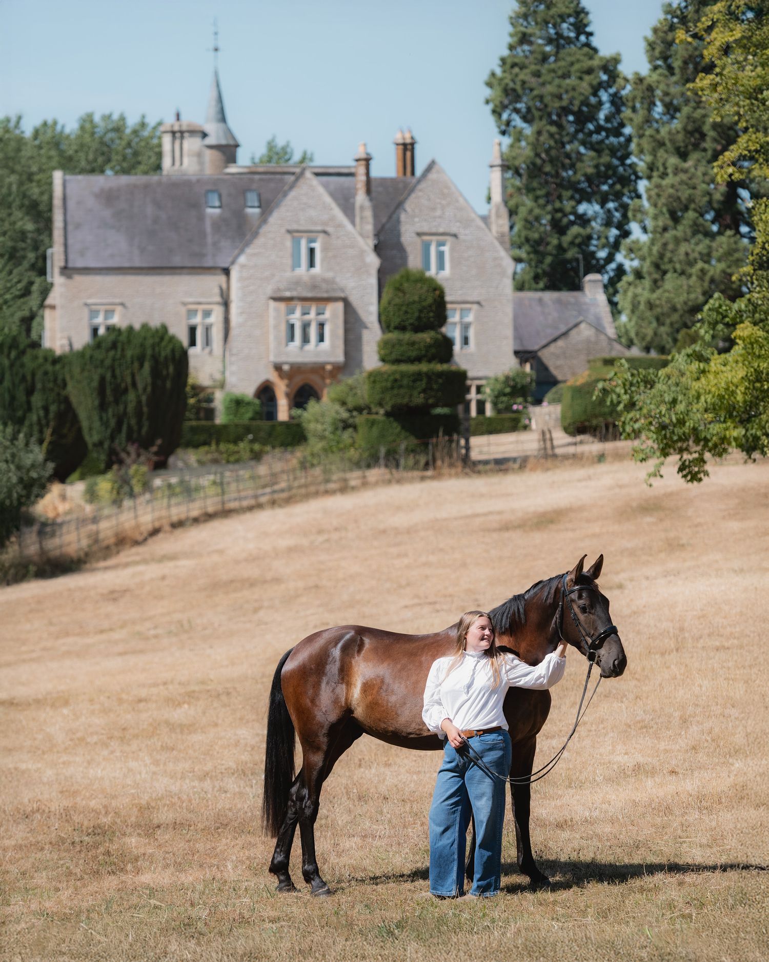 Person standing with a bay horse in a golden field before a grand stone manor house on a sunny day.