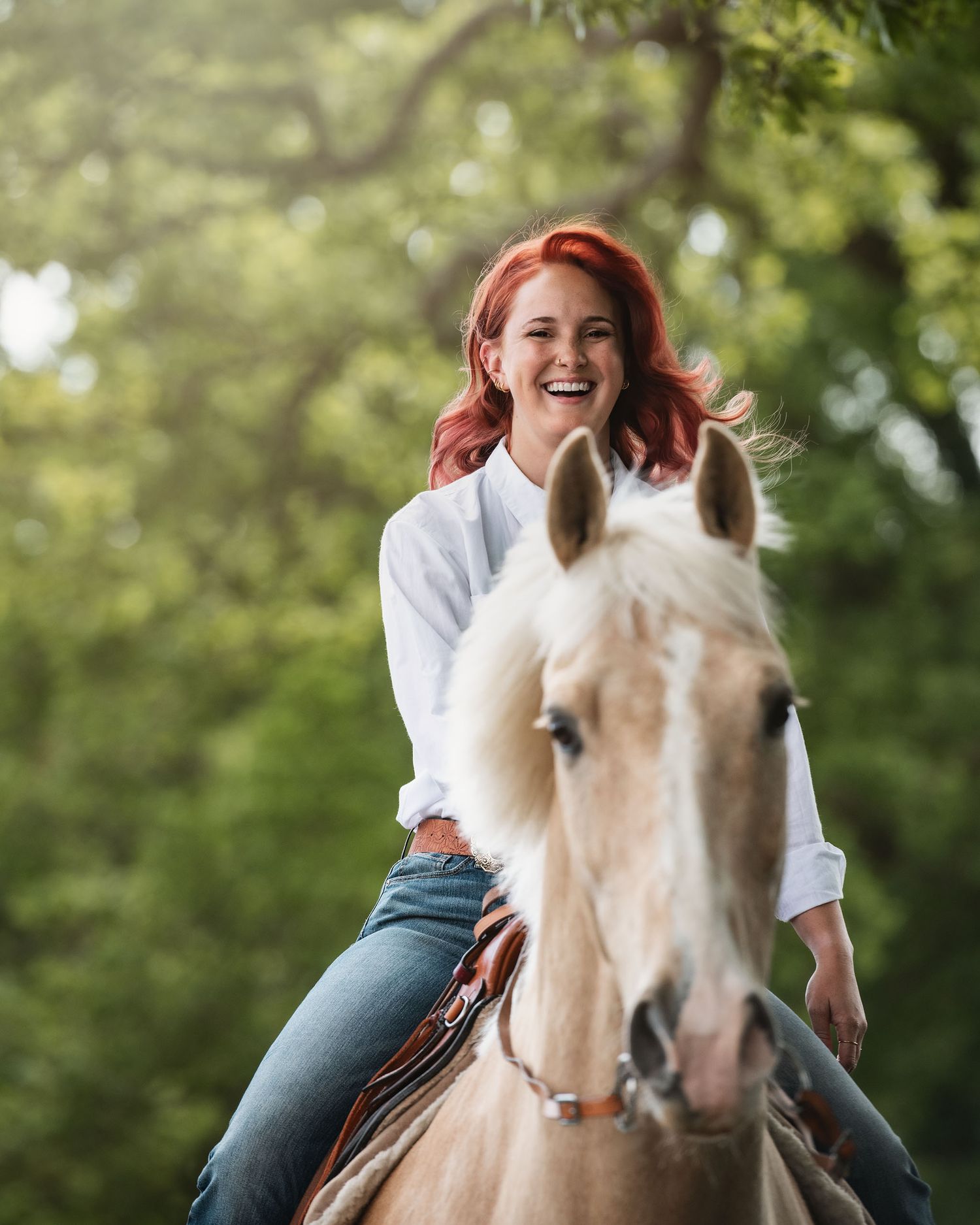 Horse and rider enjoying playful moment in sunlit forest setting. Palomino horse wearing western tack and girl with red hair.