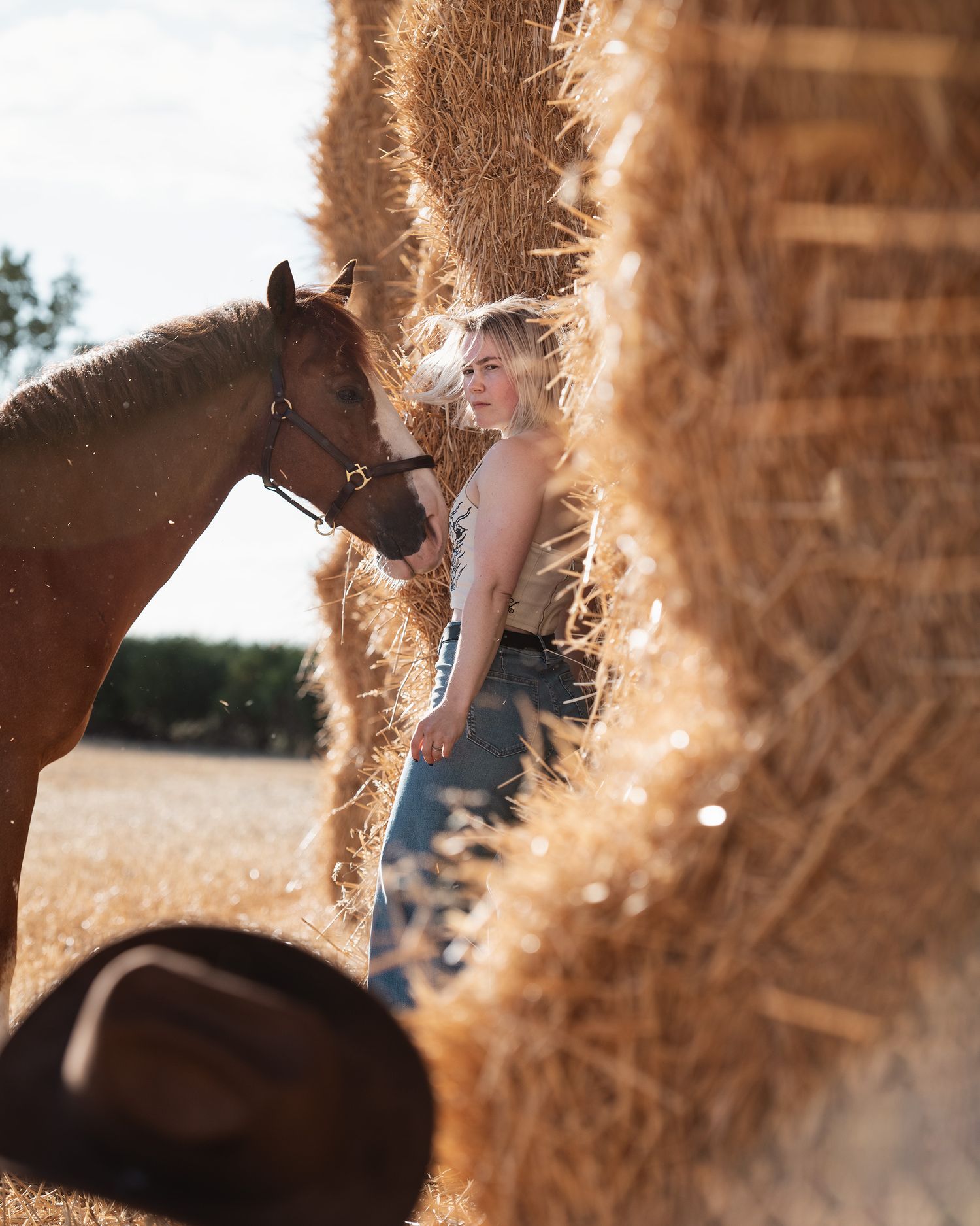 Woman in jeans stands beside a chestnut horse between hay bales on a sunny farm, with a cowboy hat in foreground.