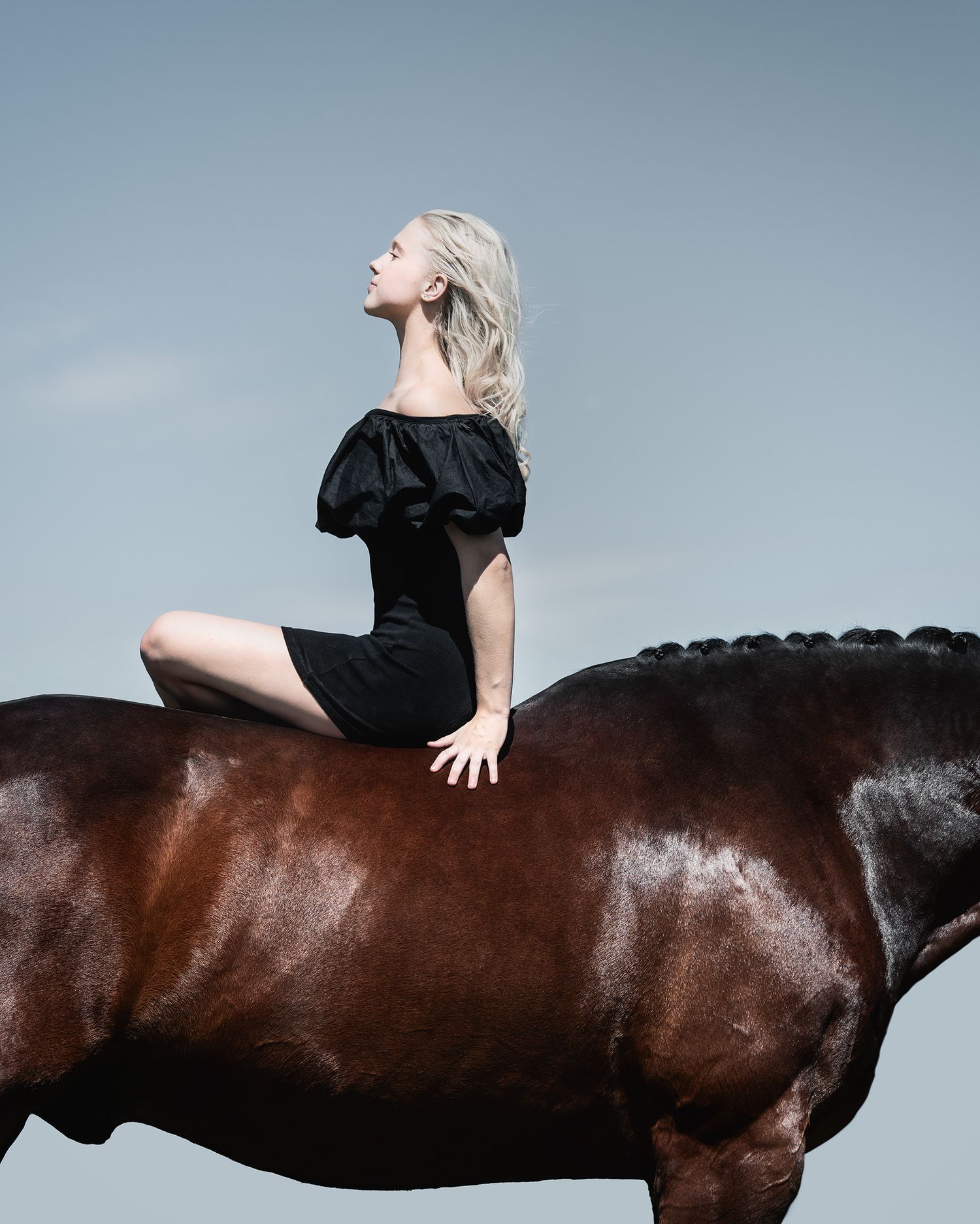 A blonde woman in a black off-shoulder dress sits gracefully atop a brown horse against a gray sky.