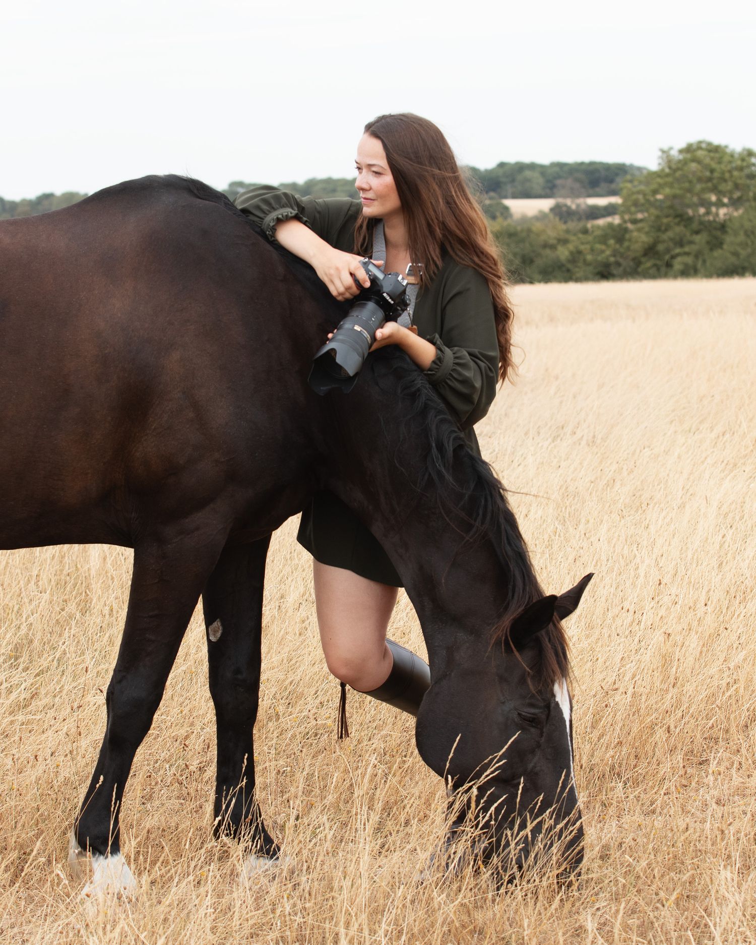 Woman in green dress grooming a black horse grazing in a golden field.