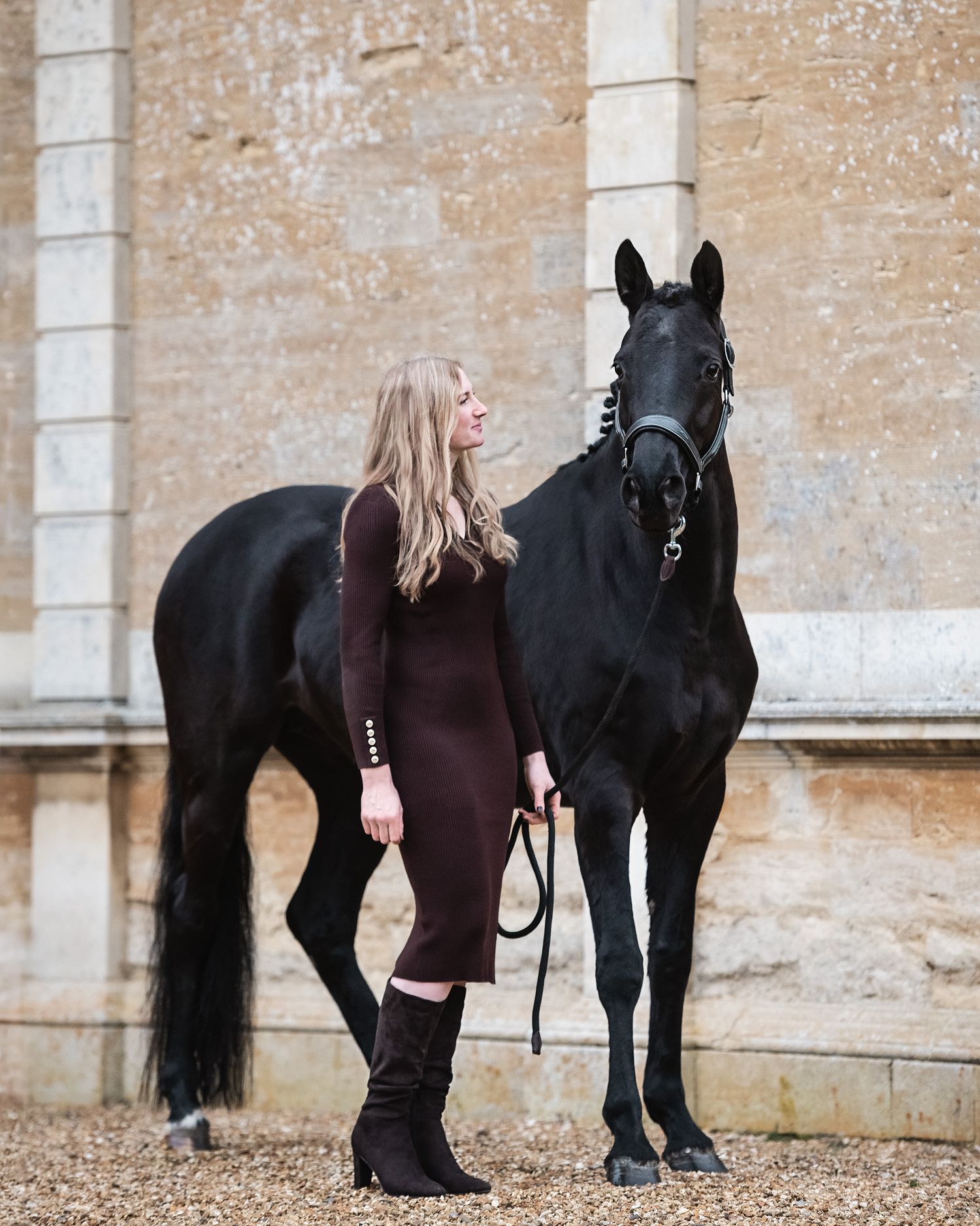 A black horse stands next to a woman dressed in Holland Cooper against a stone wall backdrop of Turvey House.