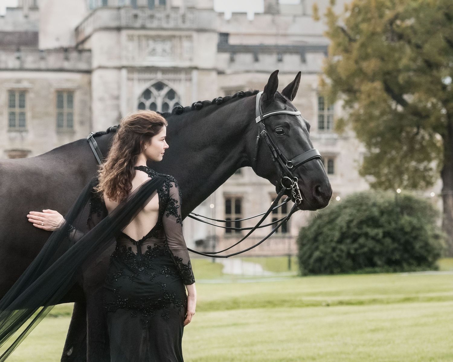 Elegant equestrian in a black dress and cape and big black horse photographed against historic castle backdrop of Ashbridge Estate.