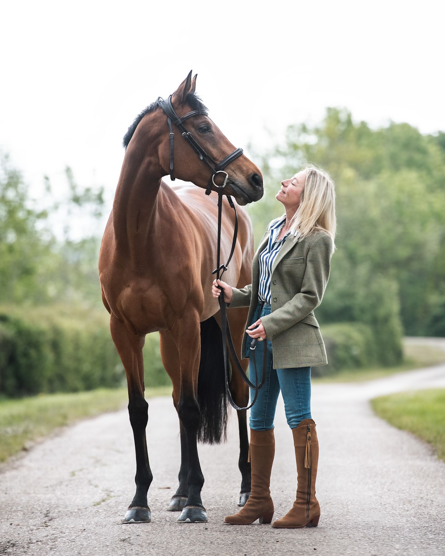 Blonde woman in tweed jacket and brown boots standing with a chestnut horse on a tree-lined path.