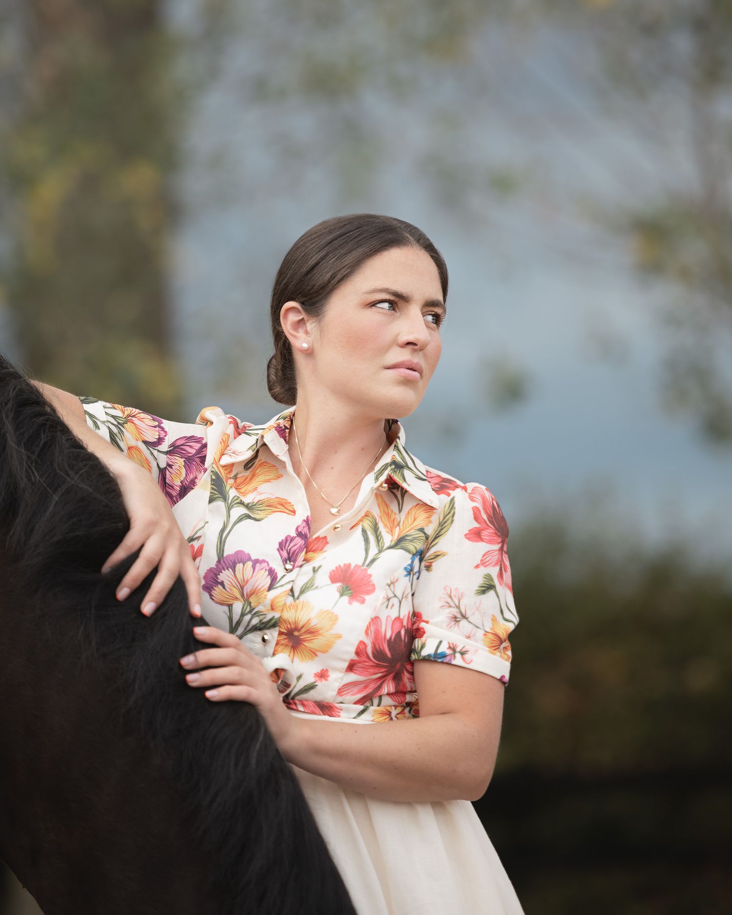 Woman in floral shirt poses beside a dark horse outdoors with soft bokeh background.