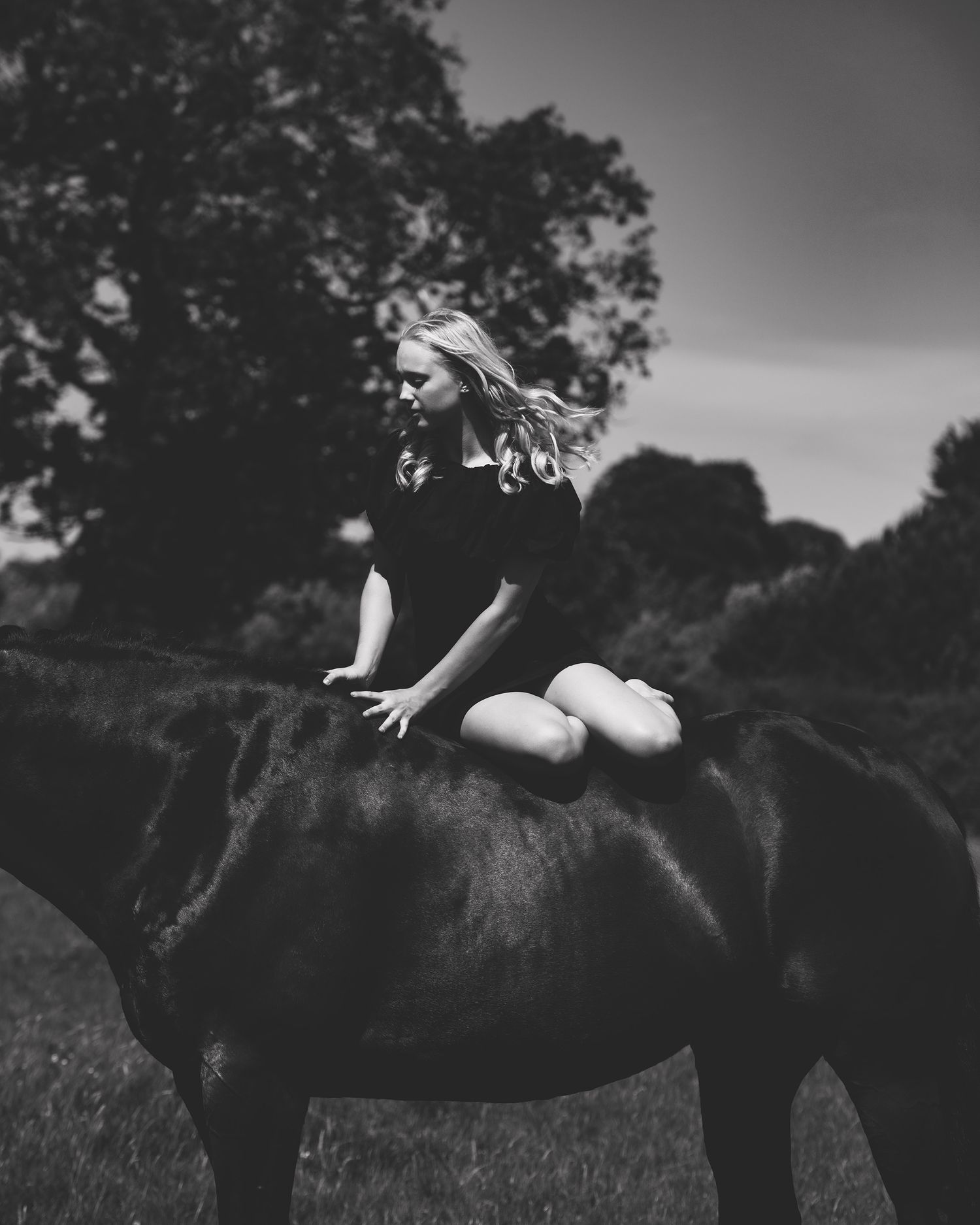Black and white photo of a young blonde girl sitting bareback on a dark horse in a field with trees.
