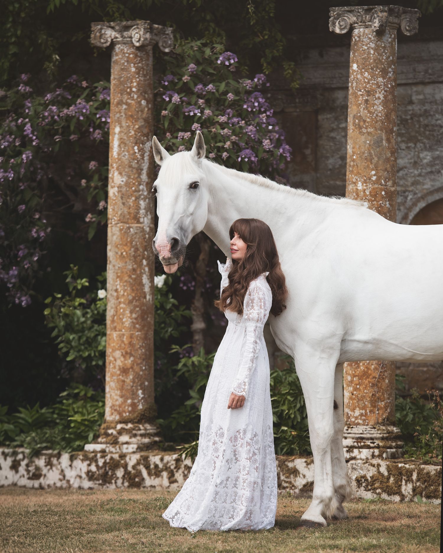 A woman in a white lace gown stands beside a white horse near stone columns with purple flowers.