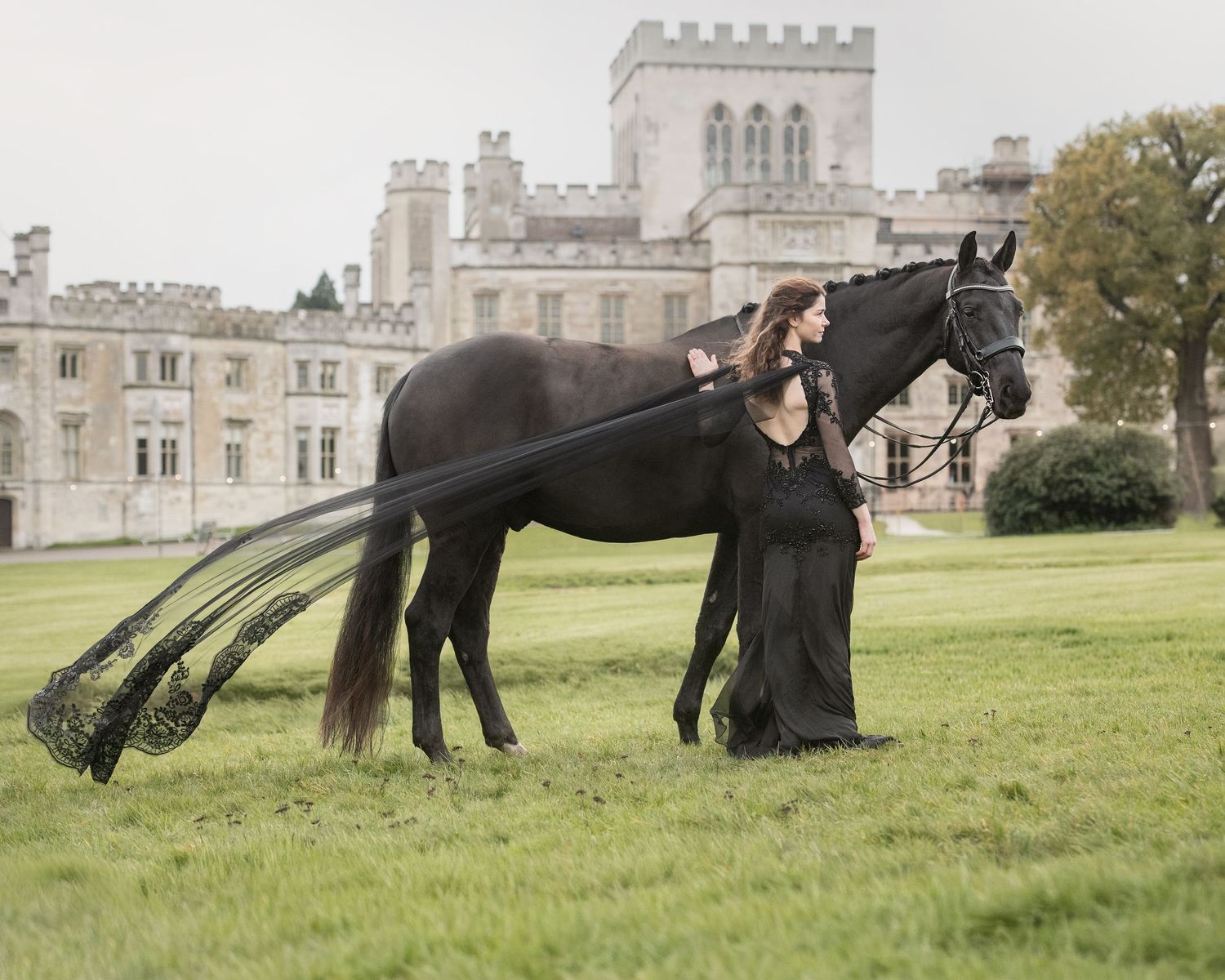 Woman in black gown stands beside a black horse with flowing tail extensions before a grand castle.
