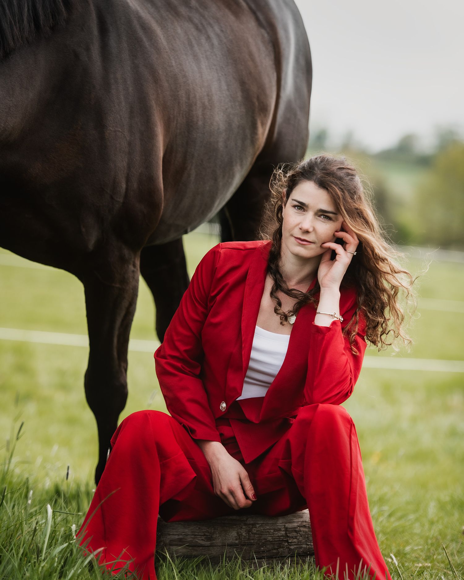 Woman in red suit sitting on a log beside a dark horse in a green field.