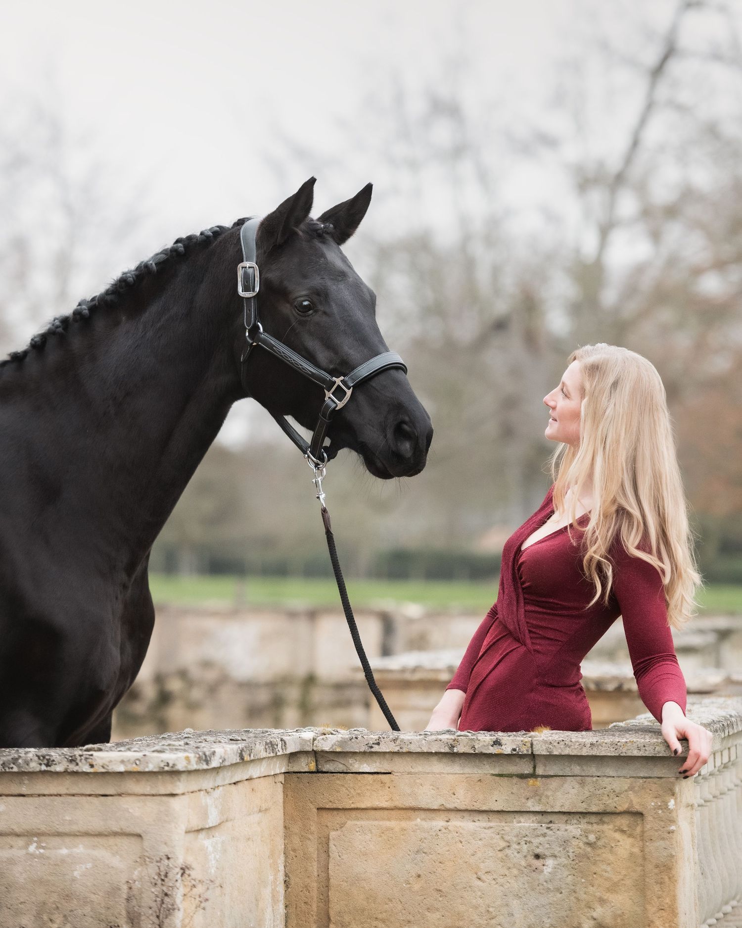 A blonde woman in a burgundy dress stands face-to-face with a black horse at a stone wall at Turvey country estate house in Bedfordshire.