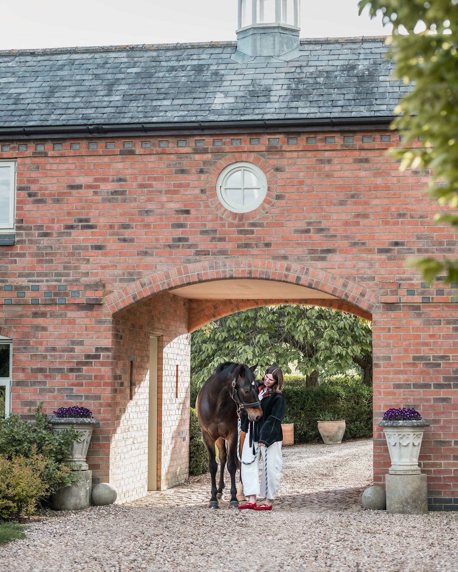 A woman stands with a dark bay horse under a red brick archway at an elegant equestrian estate.
