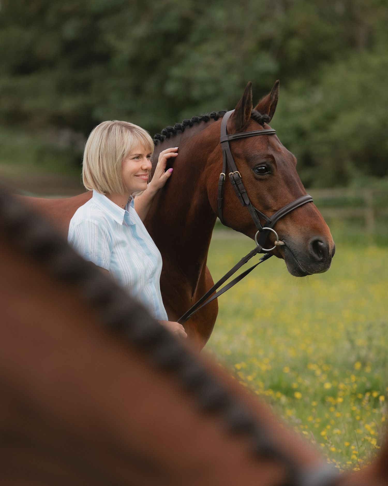 A smiling blonde woman in a light blue shirt stands beside a chestnut horse wearing a bridle in a green field.