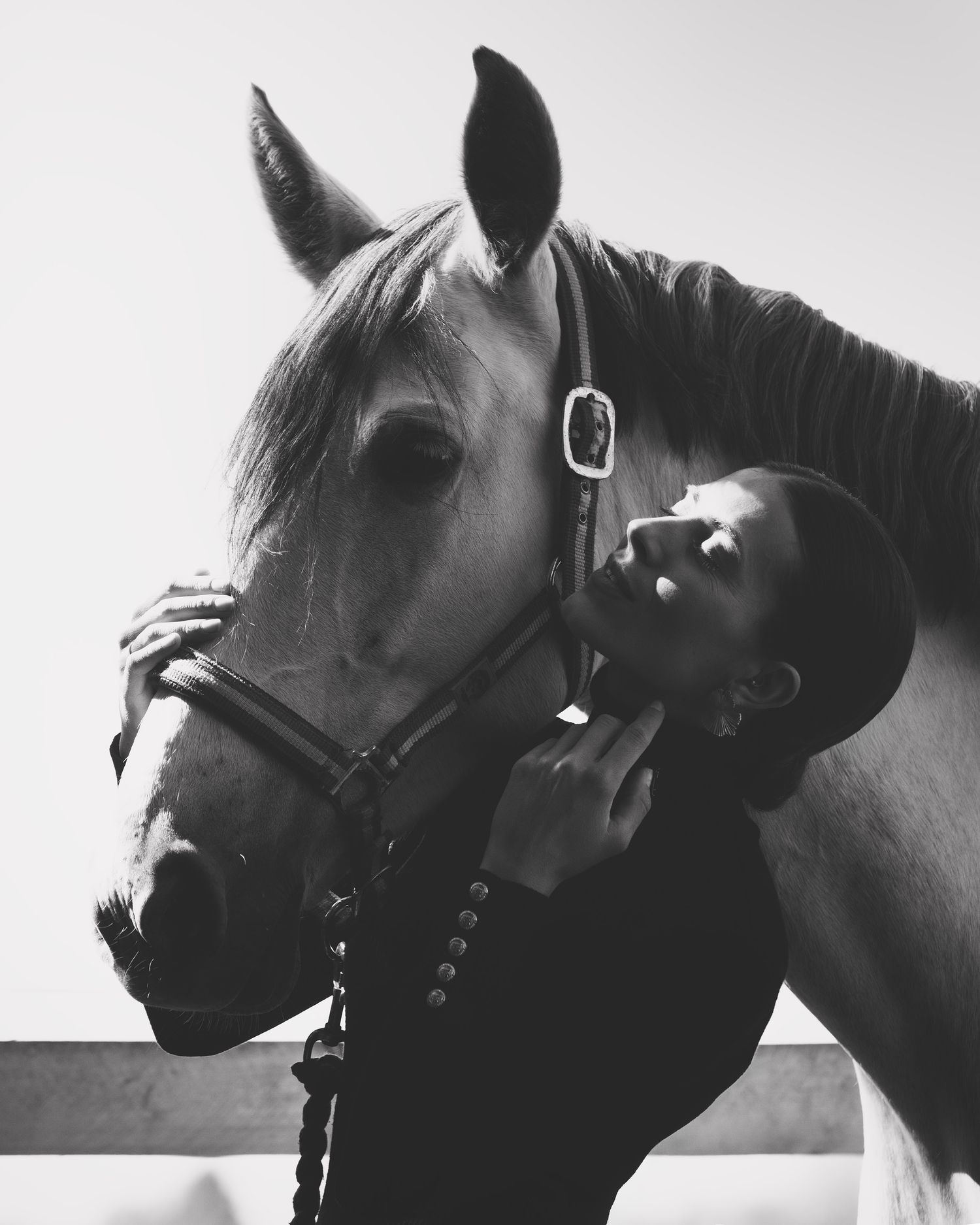 Black and white photo of a person affectionately leaning against a horse wearing a bridle outdoors.