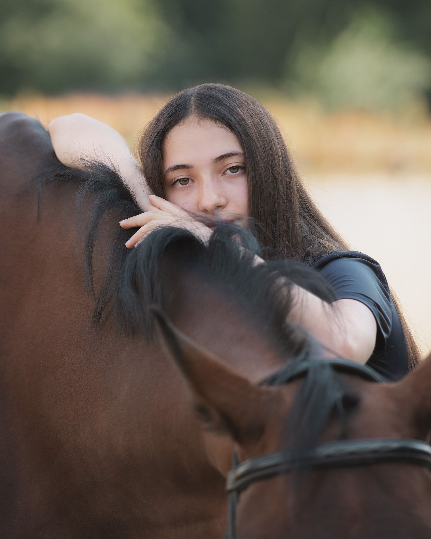 Young woman with long dark hair resting her head on a brown horse, sharing a tender moment outdoors.