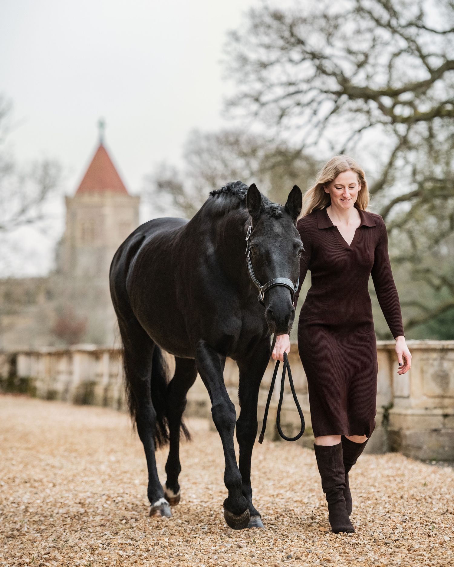 A blonde woman in a brown dress and long boots walks a black horse on a gravel path near a historic building.