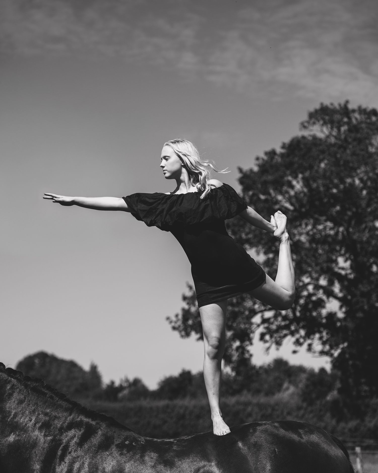 Black and white photo of a blonde woman balancing on one leg atop a horse, arms outstretched, trees in background.