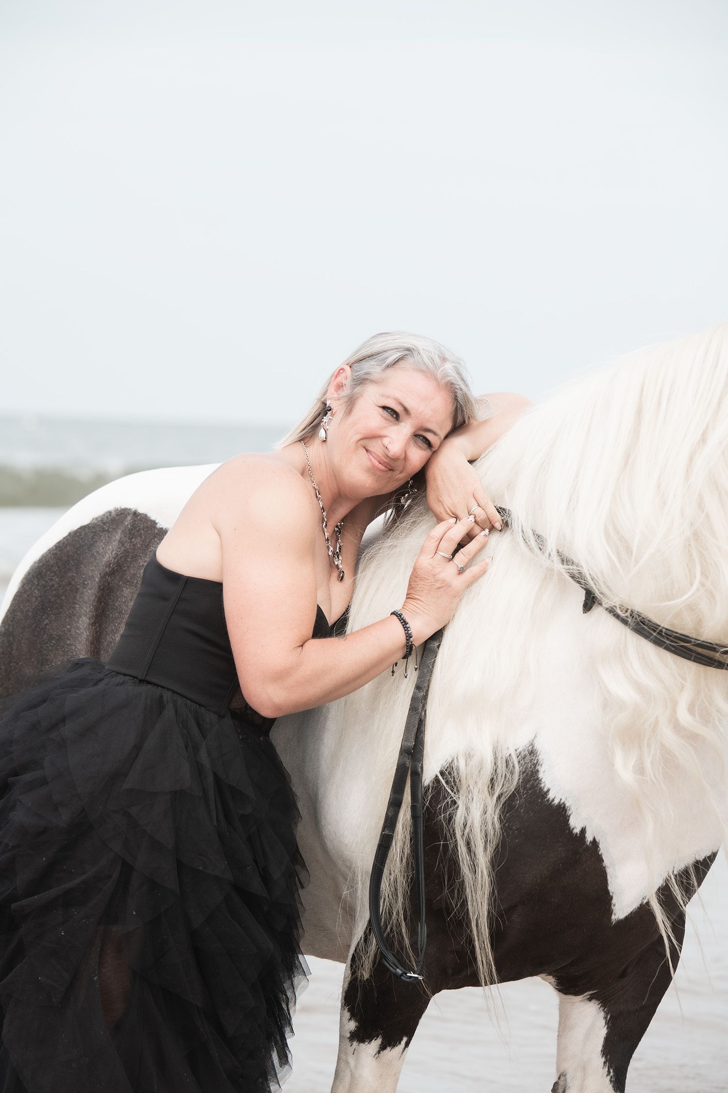 Woman in black gown embracing a white and black horse on a beach, smiling warmly.