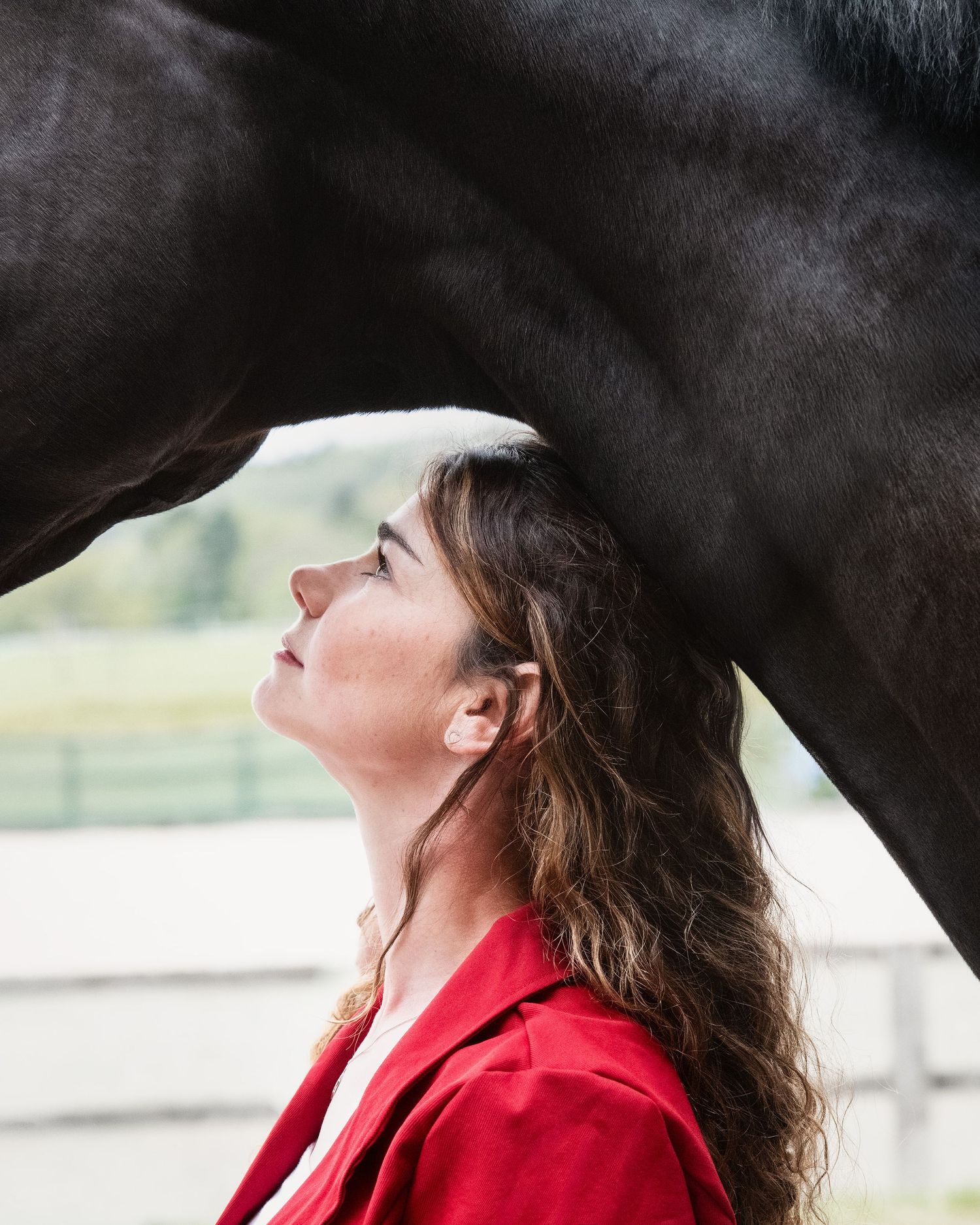 Woman in a red blazer jacket gazes upward while standing under neck of a dark black horse in a serene outdoor arena setting in Warwickshire.