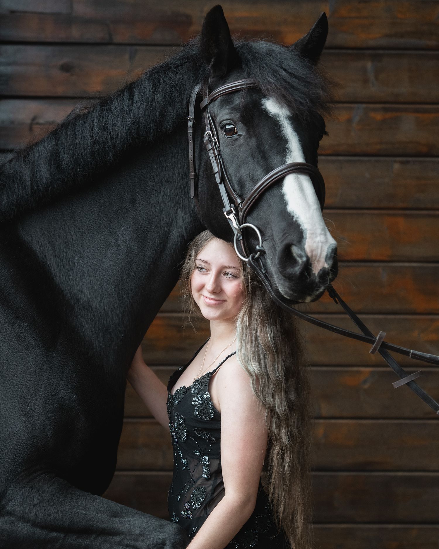 Young woman with long blonde hair in black dress poses smiling beside a black horse with white blaze in stable.