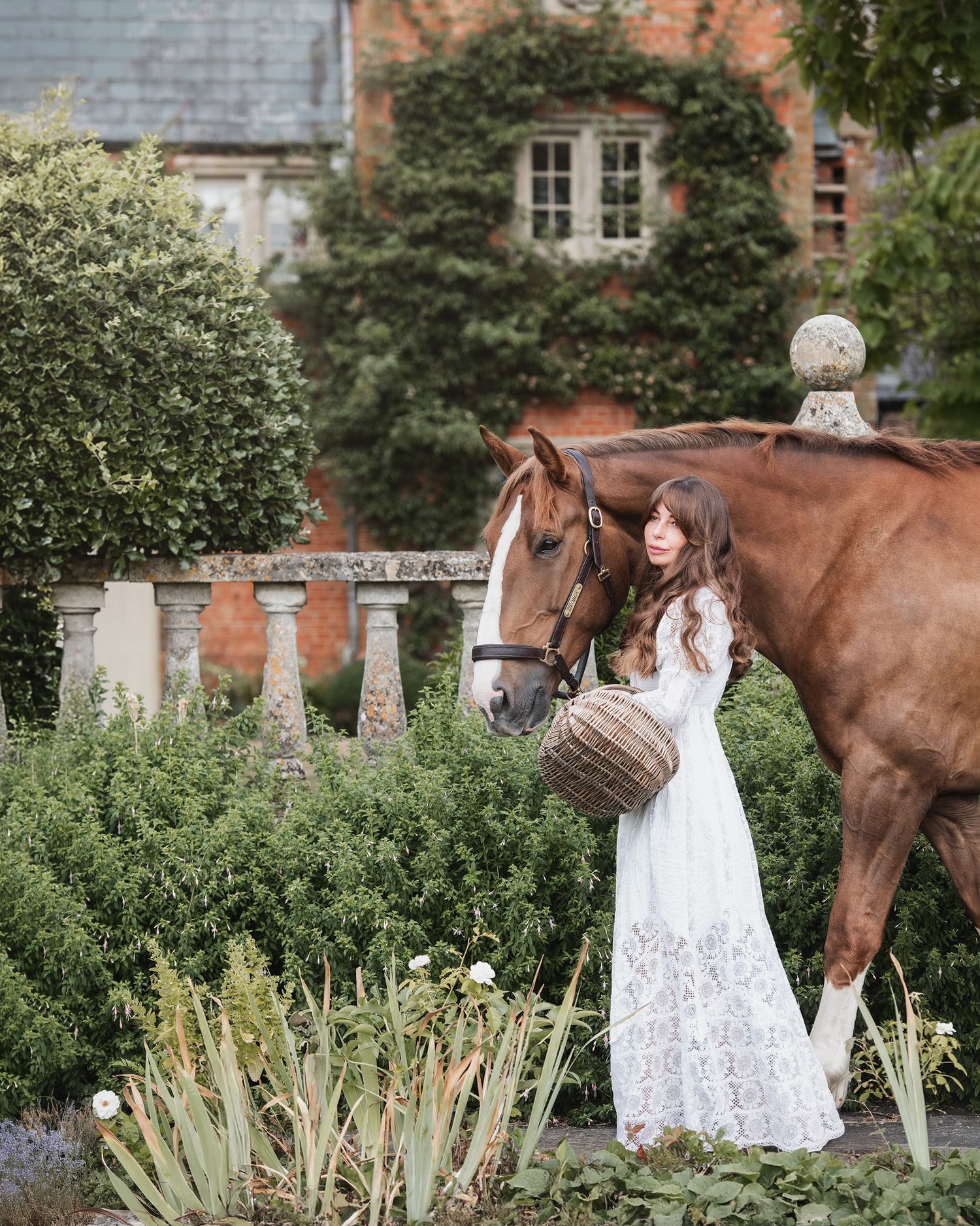 Woman in white lace dress stands beside a chestnut horse in an elegant garden with ivy-covered brick manor.