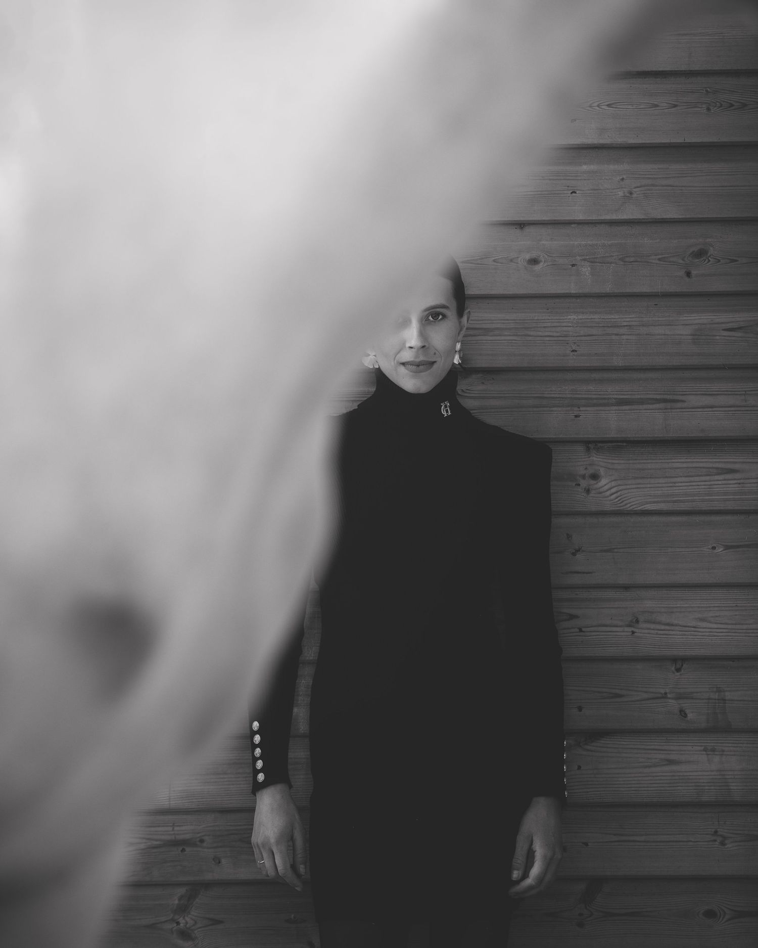 Black & white portrait of a woman in black holland cooper dress leaning against wooden wall with half horse neck, at private Stratford yard.