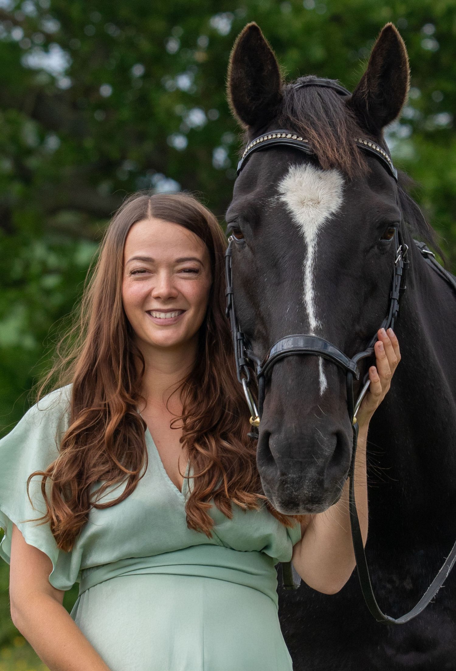 A beautiful black horse with a white blaze poses with someone wearing a mint green top against a leafy background.