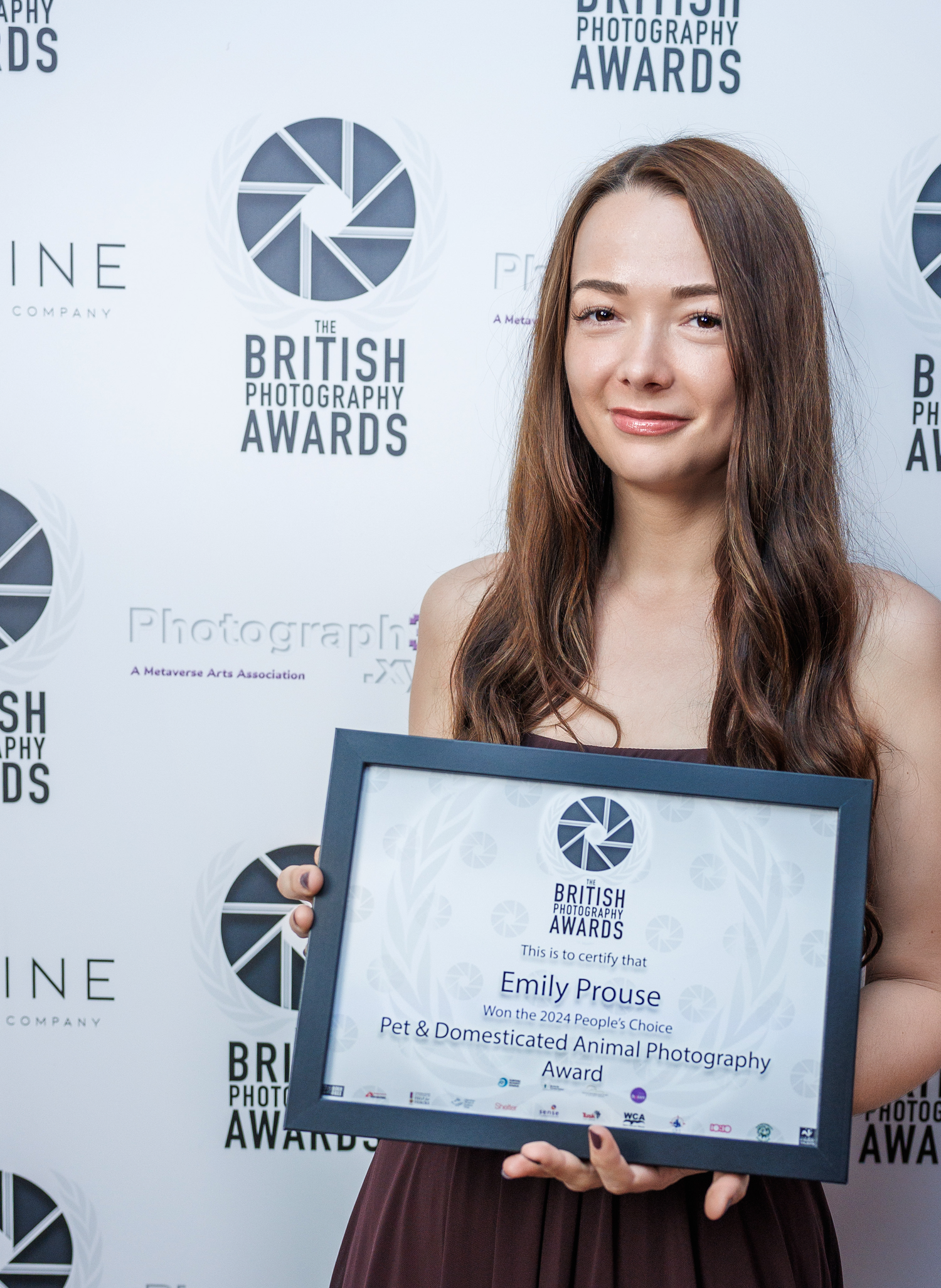Award recipient holding photography award certificate at British Photography Awards ceremony with logo backdrop.