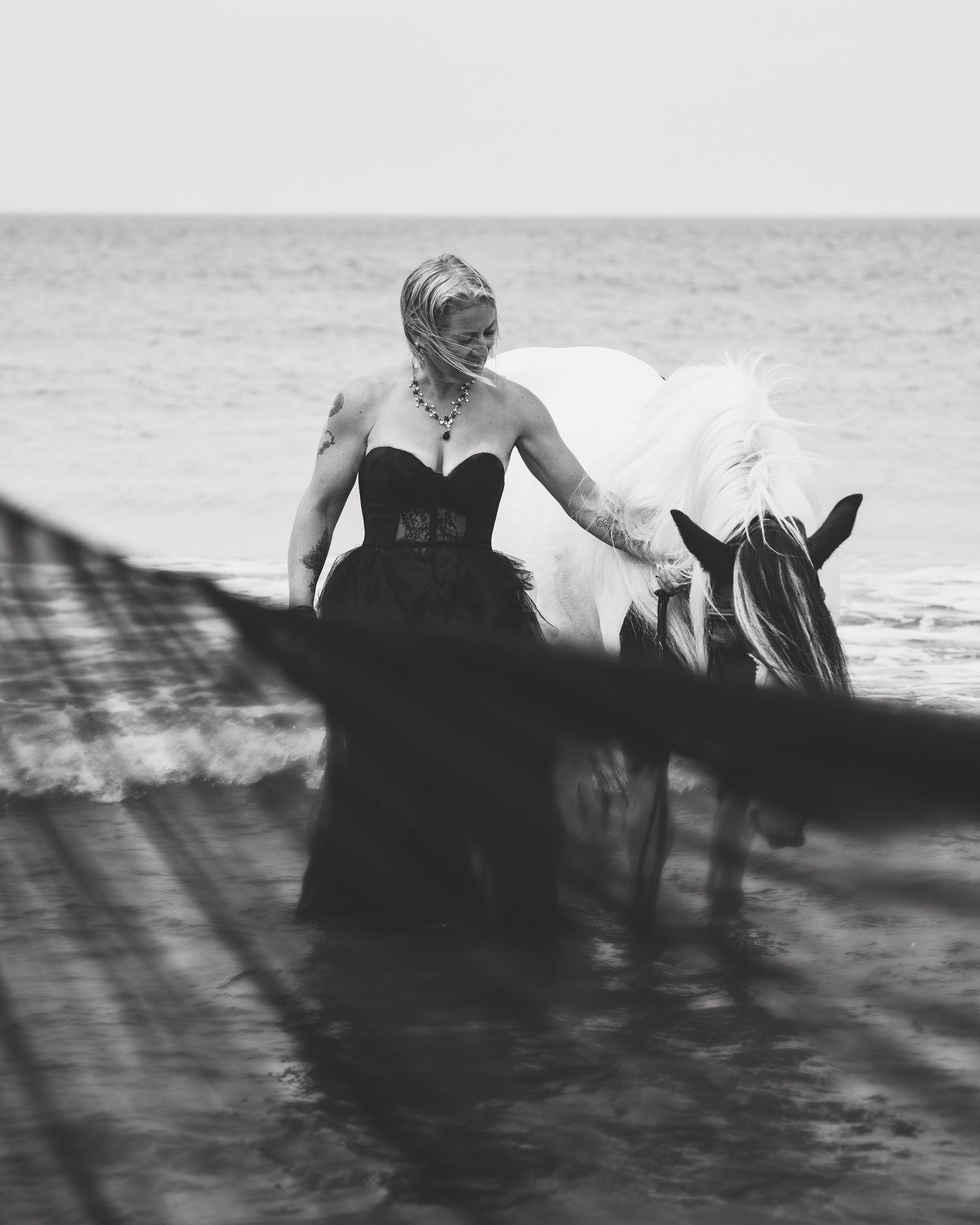Woman in black gown stands with white horse in ocean waves, black and white photography.
