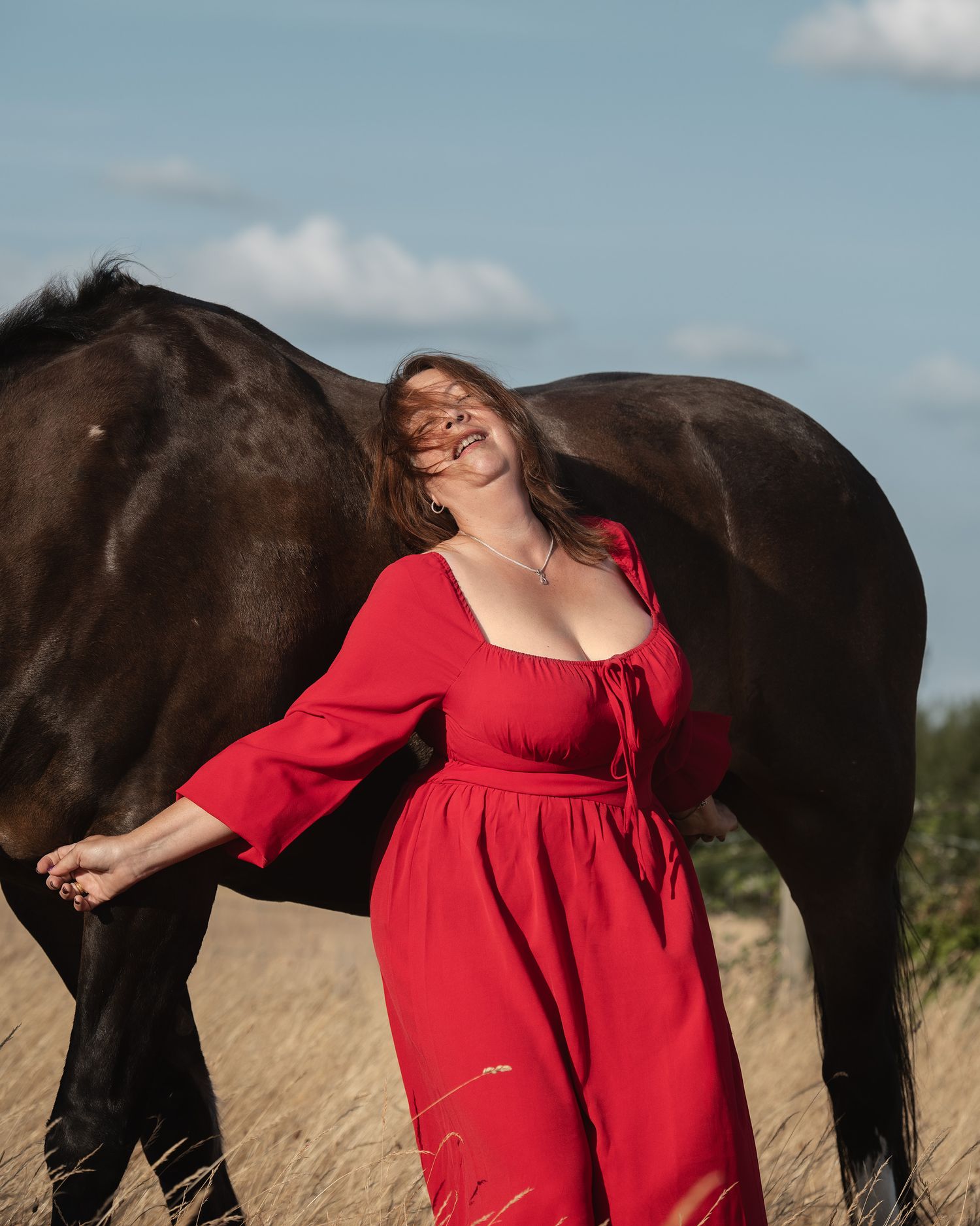 Woman in red dress standing beside a dark horse in a golden field under a blue sky.