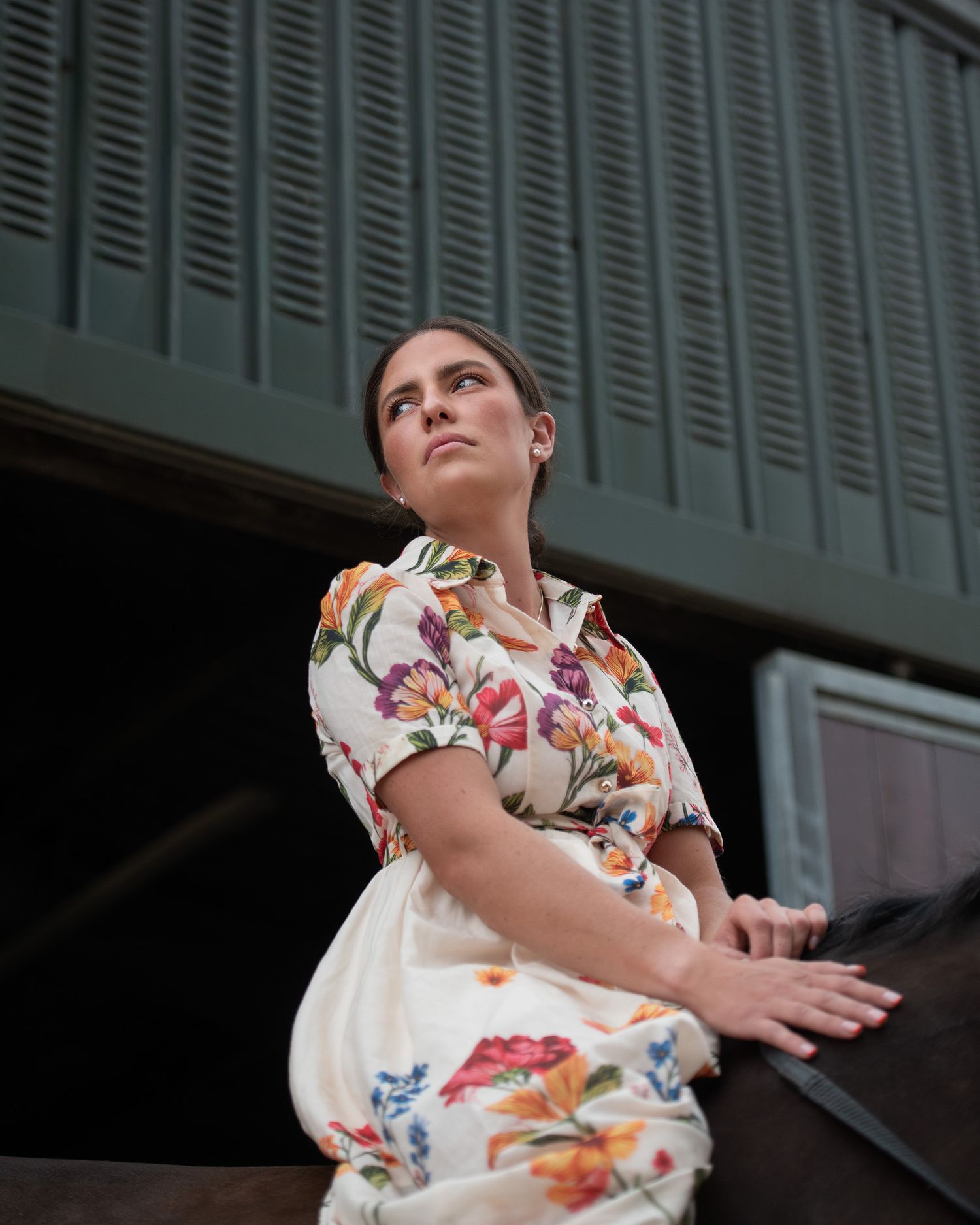 Woman in floral dress sitting outdoors, gazing upward near a green building.