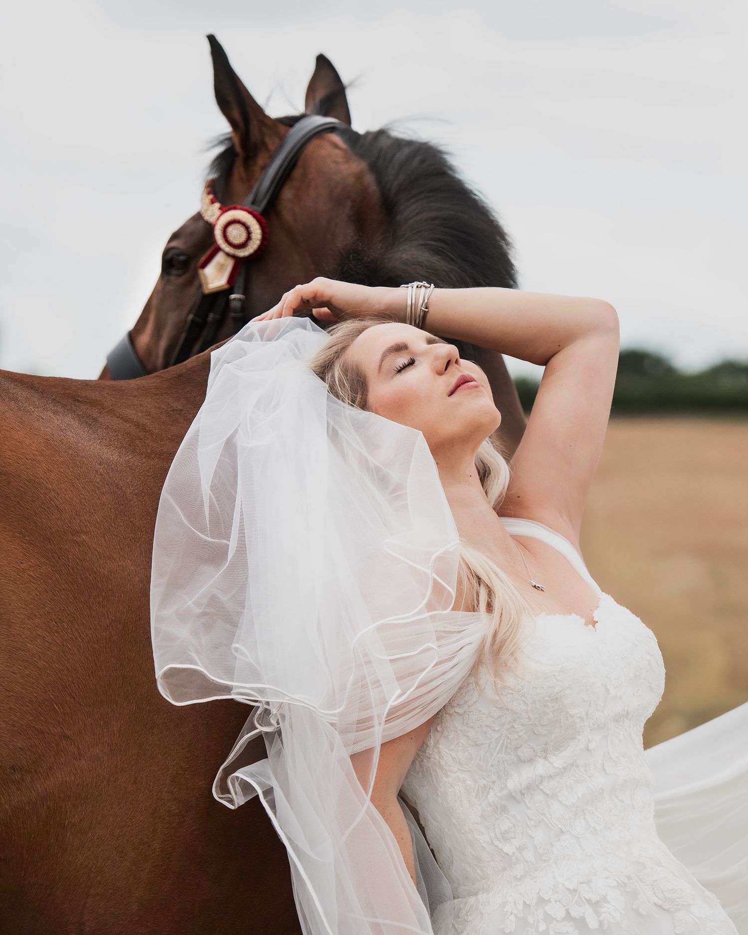 Bride in white lace gown and veil leans against a chestnut horse wearing a rosette in an outdoor field setting.
