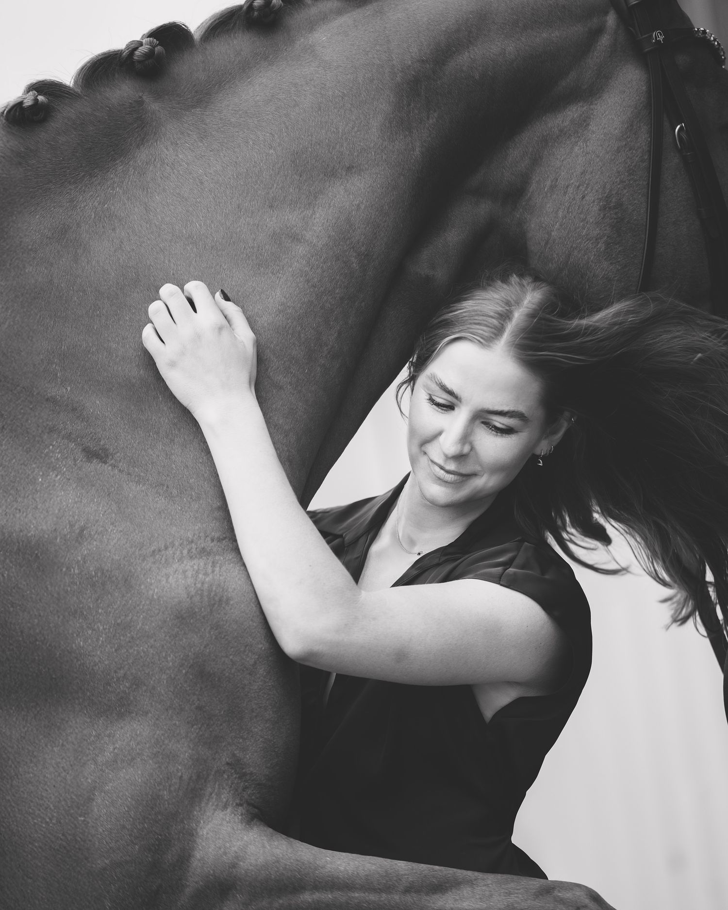 Black and white photo of a smiling woman leaning against a dark horse, her hair flowing as she touches its side.