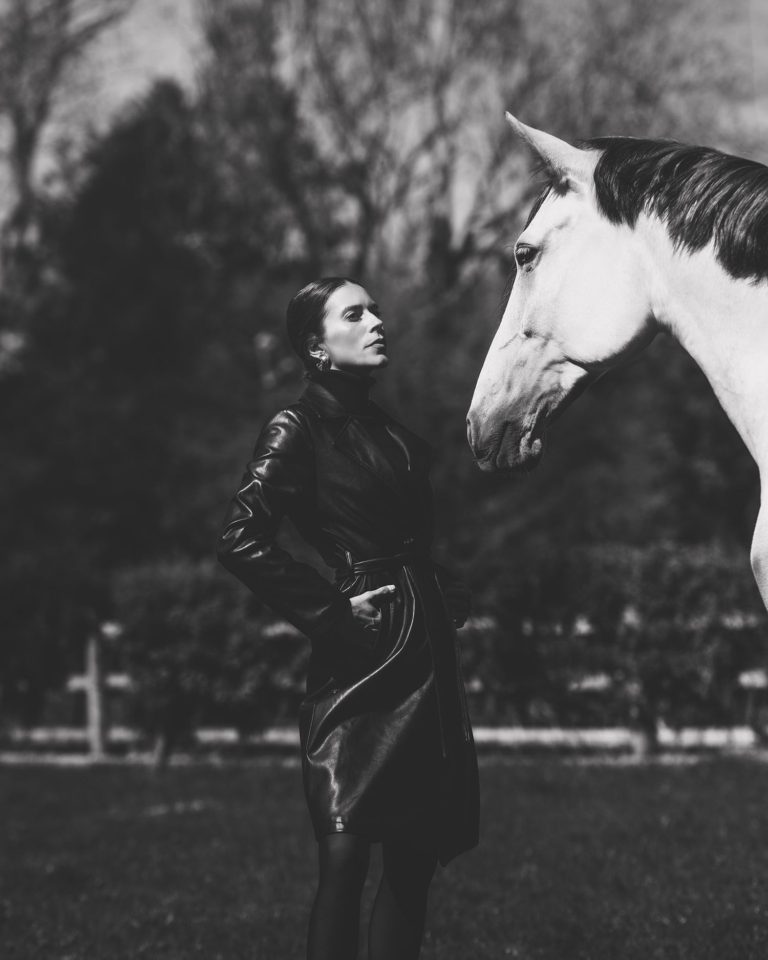 Black and white photo of a woman in leather jacket standing beside a white horse in an outdoor field.