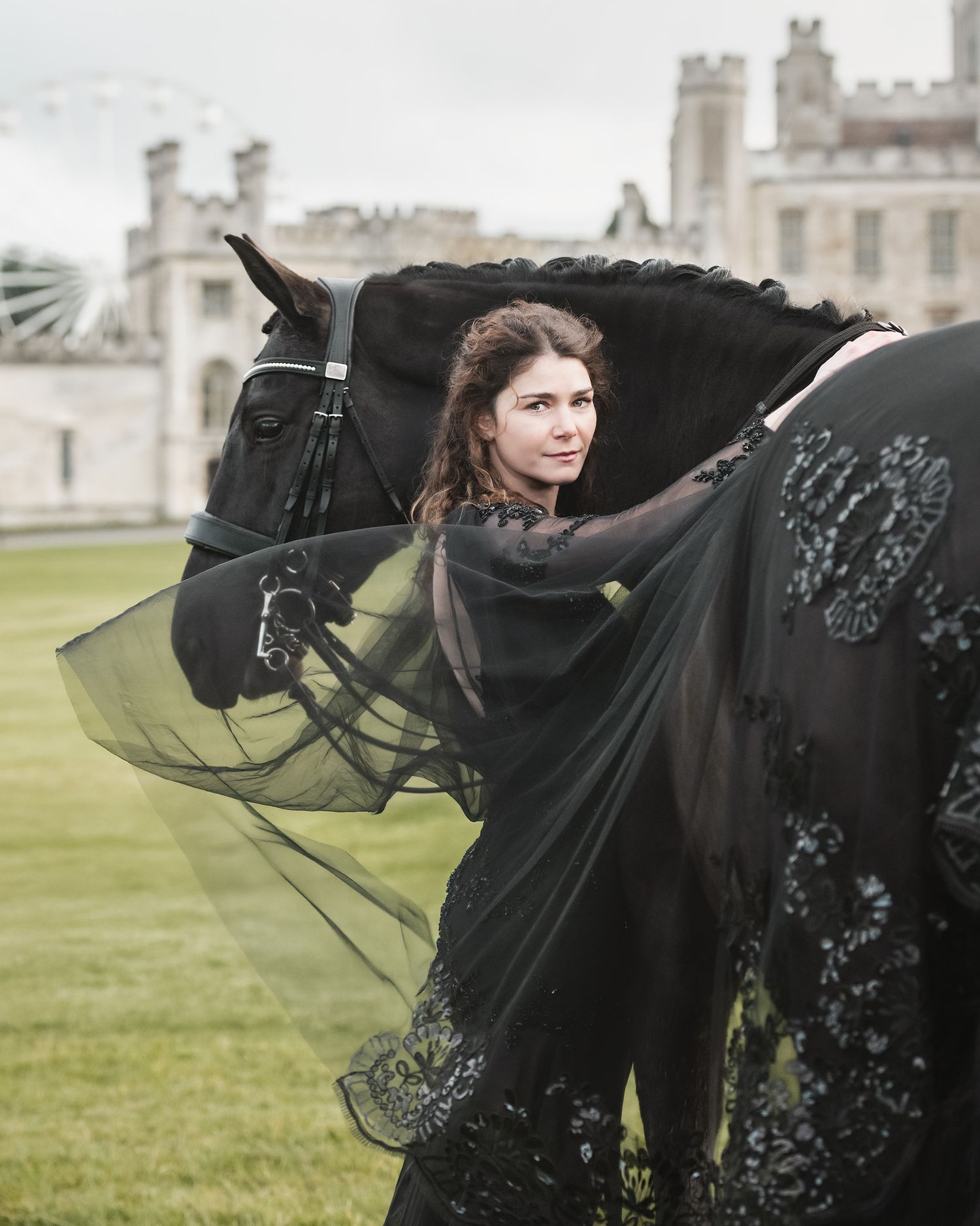 A dressage horse with dressage rider trainer in a black flowing gown against a misty outdoor castle estate house backdrop.