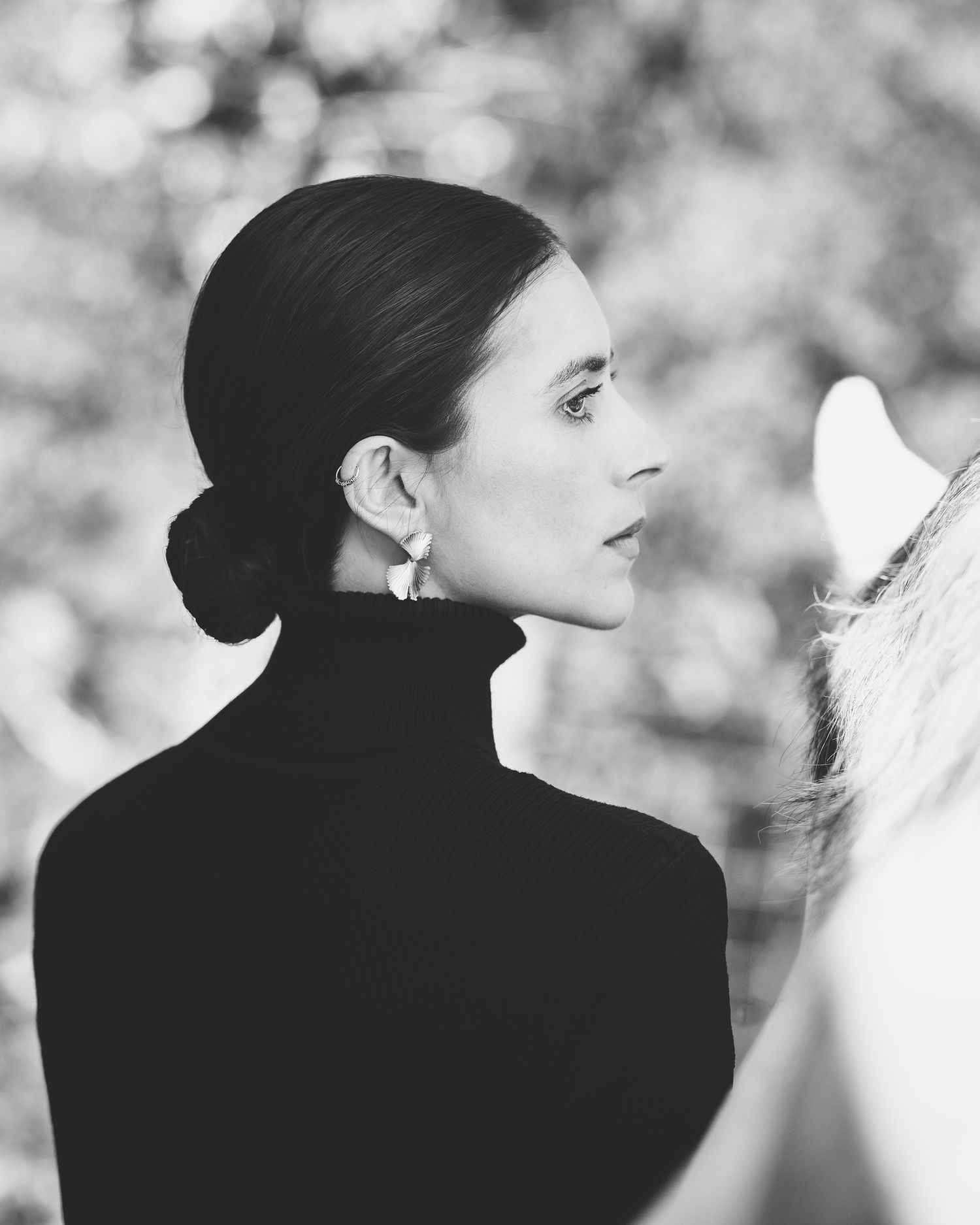 Black and white profile portrait of elegant woman with sleek bun, pearl earring, black turtleneck, beside white horse.