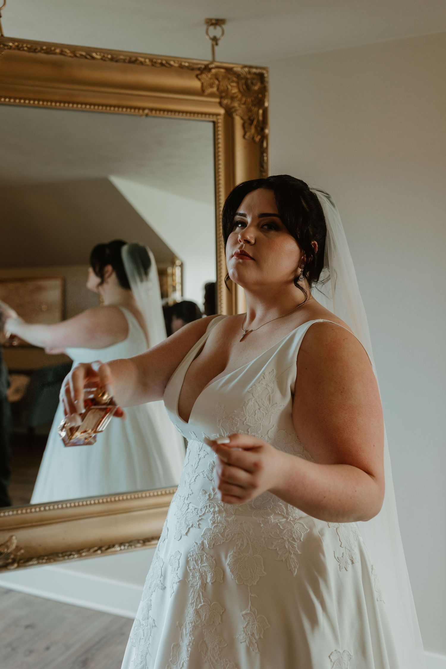 Bride in elegant white dress looking in ornate gold mirror during wedding preparations.
