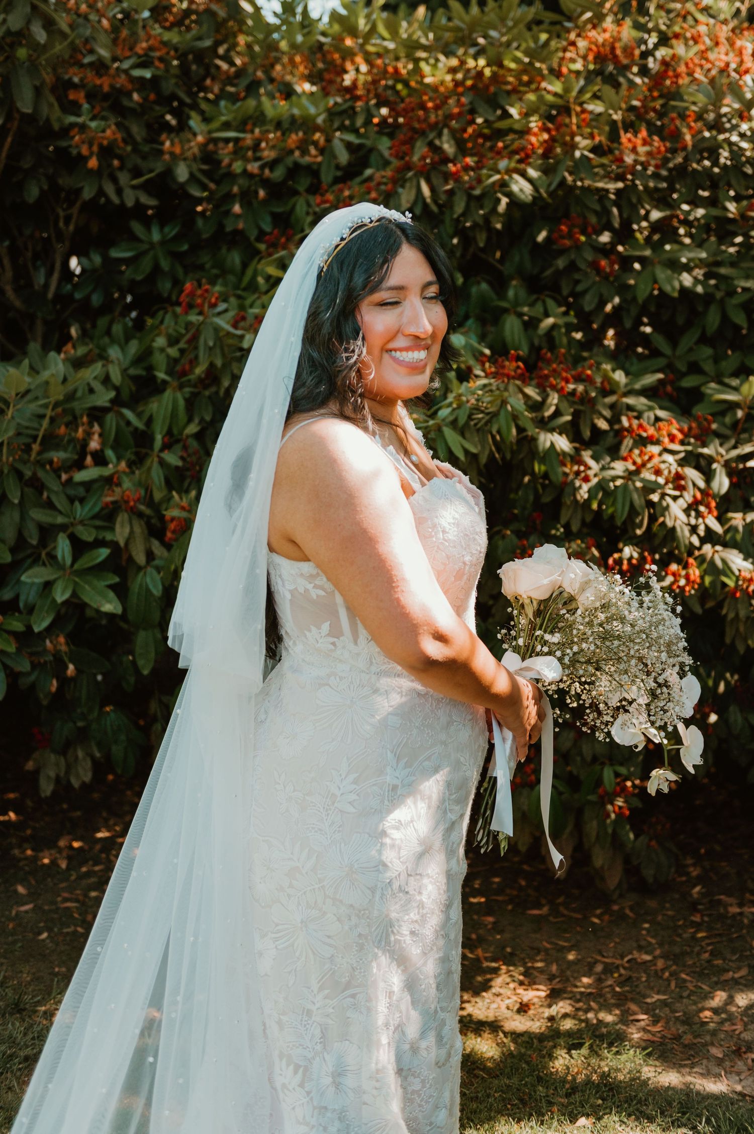 Radiant bride in lace dress with flowing veil poses against flowering garden backdrop.